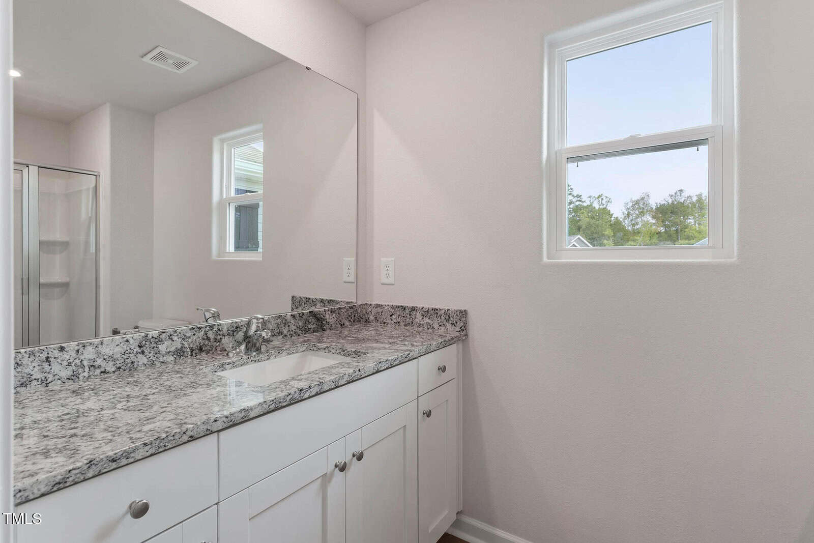 White bathroom with a granite countertop featuring a single sink. A large mirror above the vanity reflects the room, and there are two windows allowing natural light. A glimpse of a glass-enclosed shower is visible on the left.