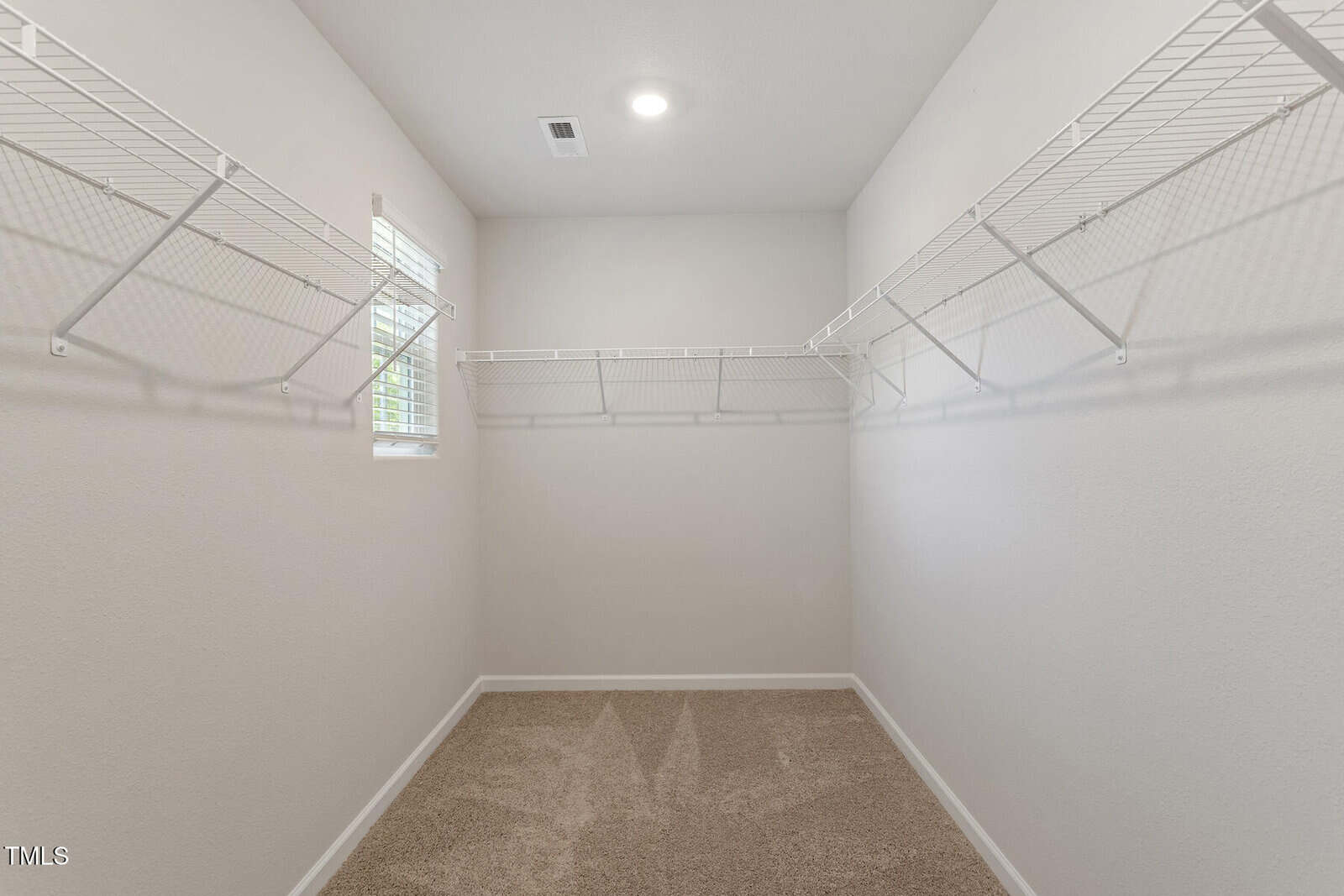 Empty walk-in closet with beige carpet and white walls, featuring wire shelving on three sides. A small window on the left lets in natural light, and a ceiling light is centered above.