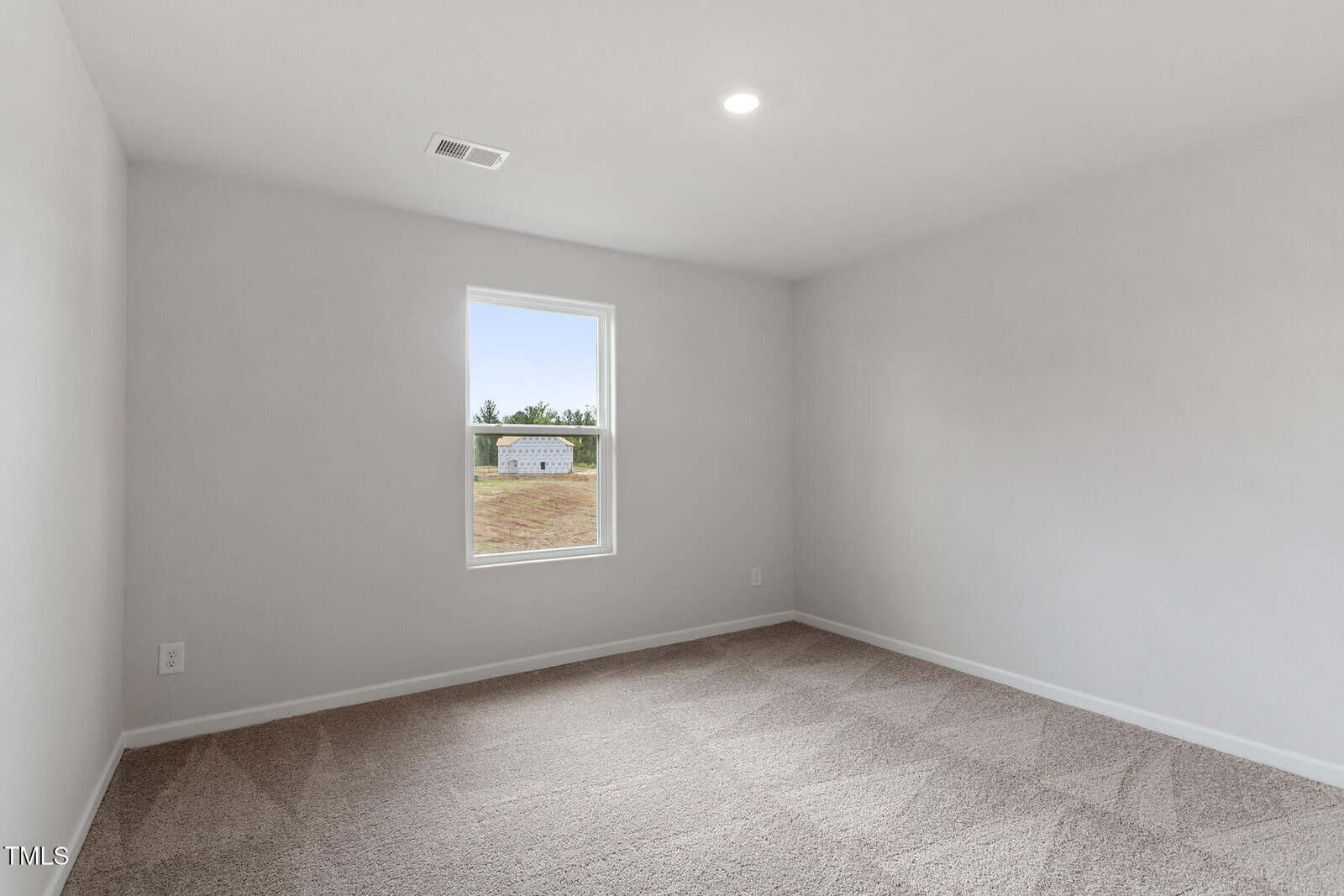 A vacant room with light gray walls and a carpeted floor. A single window on the far wall shows a view of a small building and greenery outside. The ceiling has a recessed light fixture.