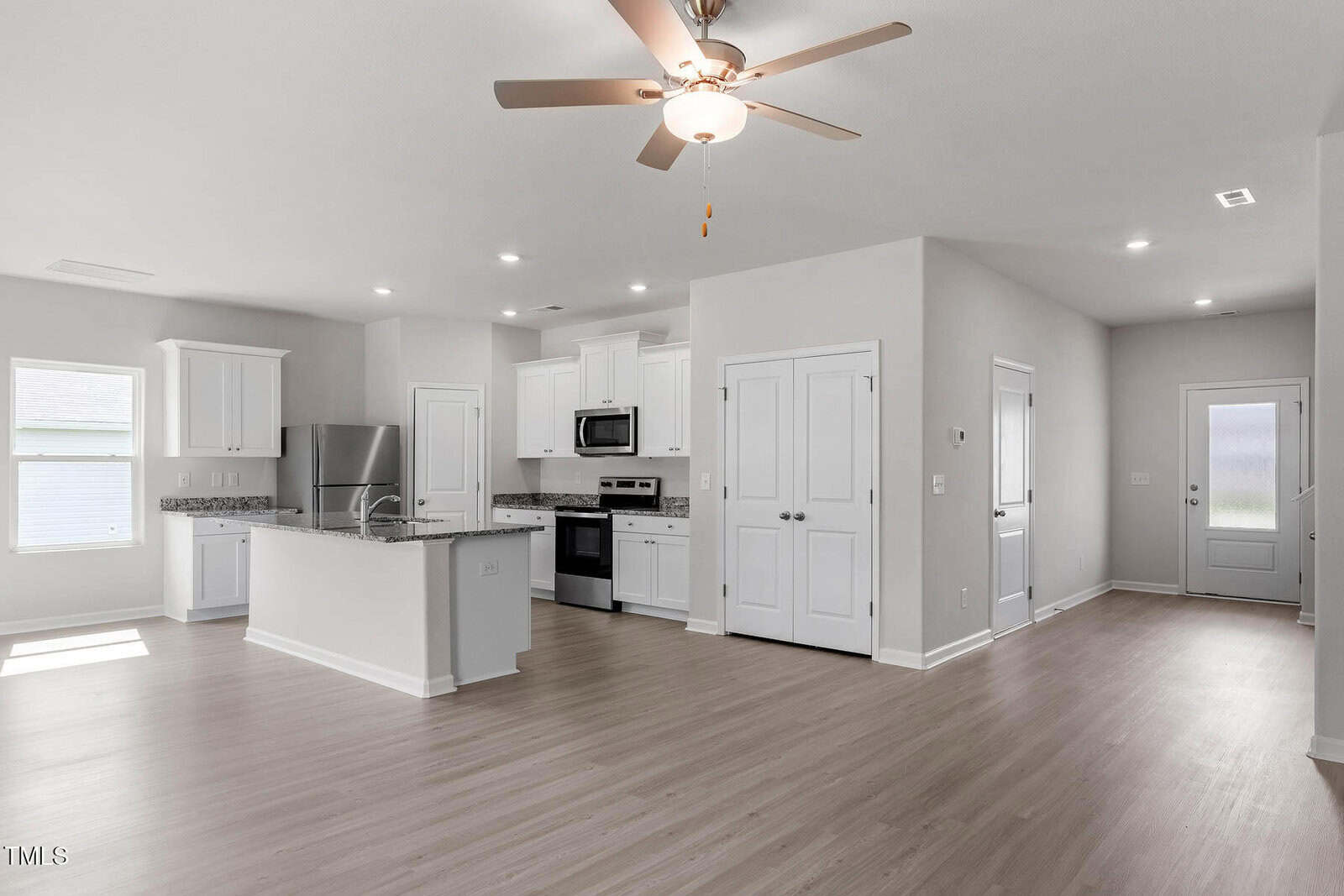 Open-concept kitchen and living area with light wood flooring. The kitchen features white cabinets, a central island with granite countertops, stainless steel appliances, and a ceiling fan. A door leads to an adjacent hallway.