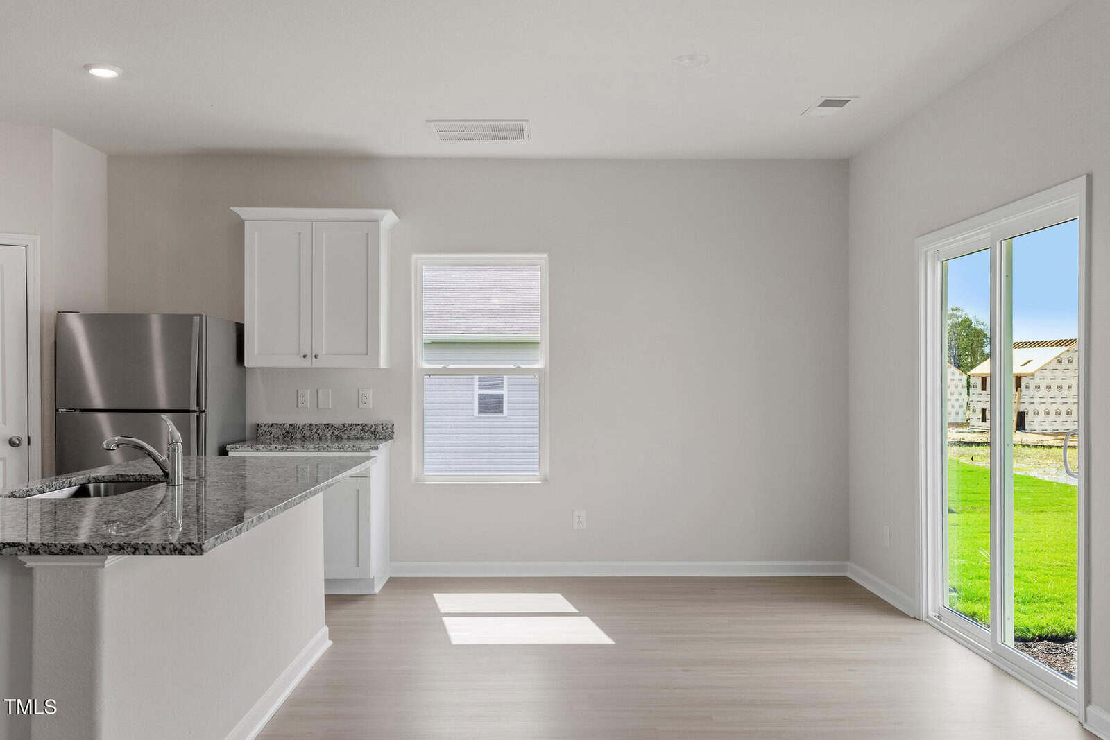 A bright, empty kitchen and dining area with light wooden flooring. The kitchen features white cabinets, a granite countertop, and a stainless steel refrigerator. A sliding glass door on the right opens to a grassy backyard with partial fencing.