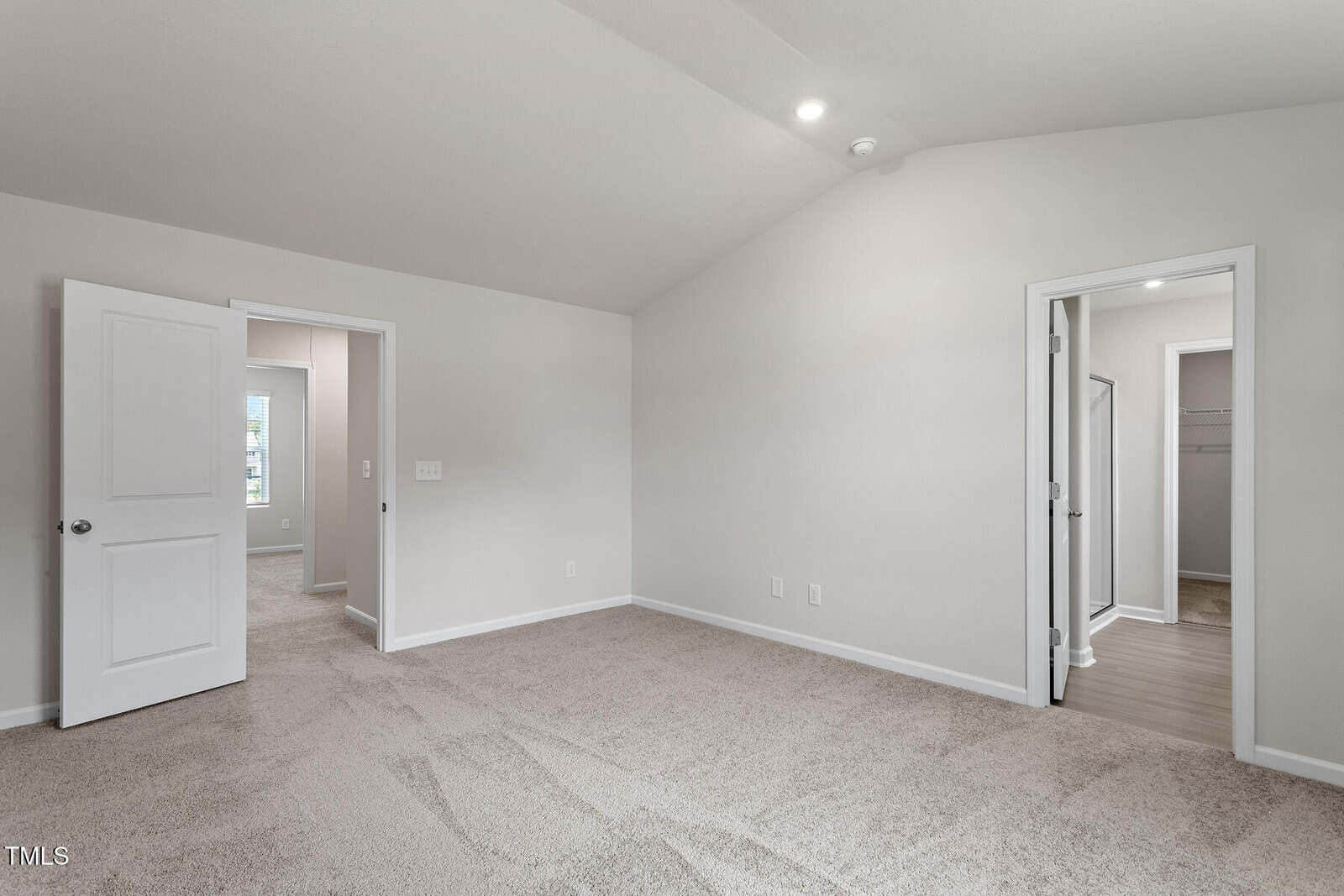 Empty carpeted room with beige walls and a vaulted ceiling. An open door on the left leads to another room. On the right, there is an open doorway leading to a small hallway and a closet. Two recessed lights are on the ceiling.