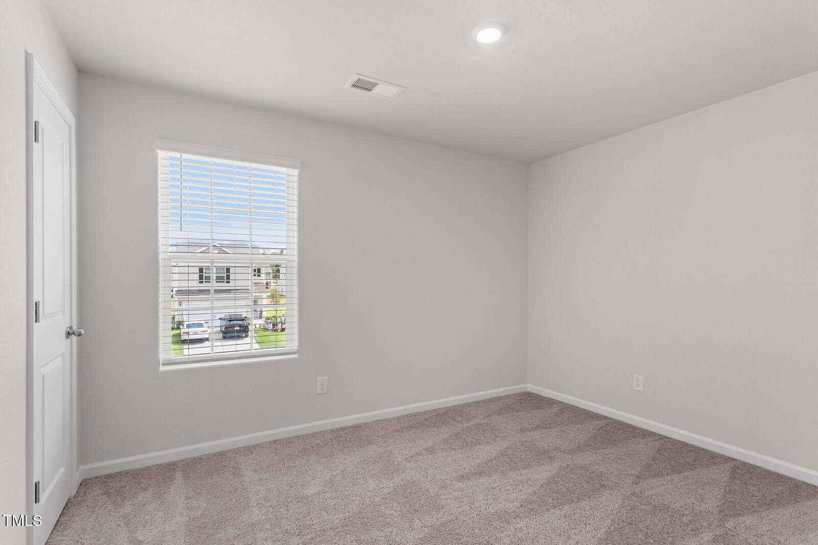 Empty, carpeted room with beige walls and a white ceiling. A single window with blinds is on the left wall, and a white door is visible. The room is lit by a ceiling light, and sunlight enters through the window.
