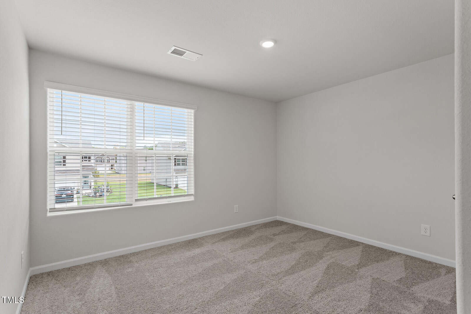 Empty room with beige carpet and white walls. A large window allows natural light, showing a view of neighboring houses. The ceiling has a single recessed light, and there's a visible wall outlet.