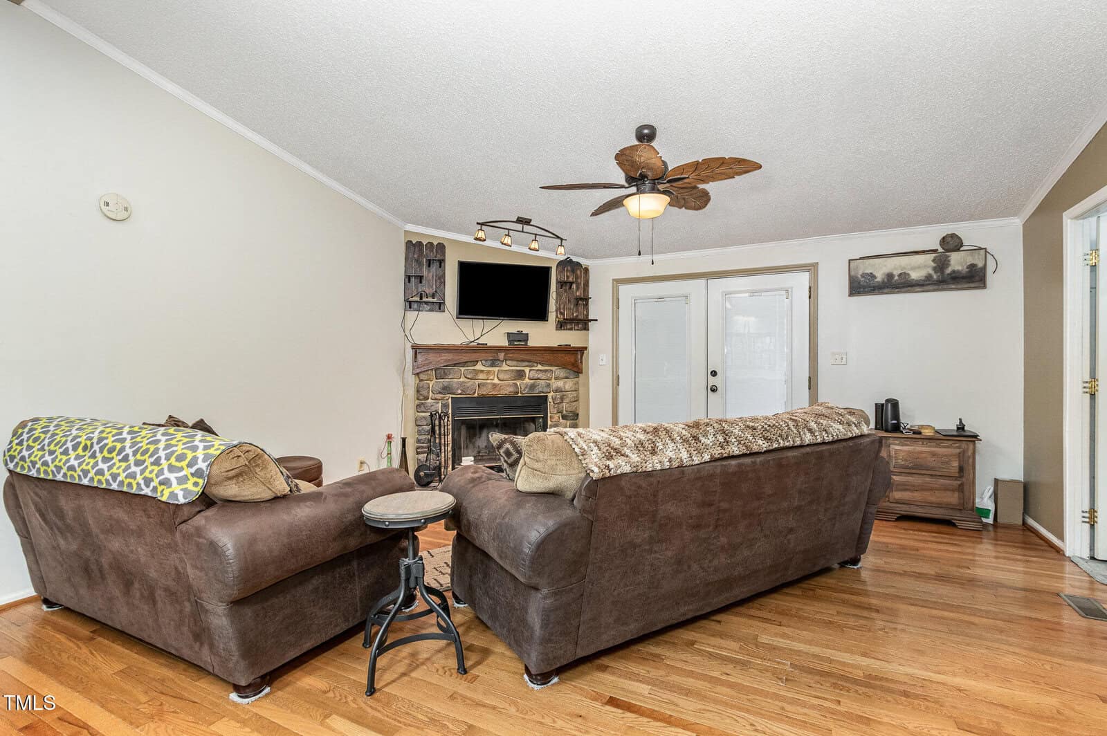A living room features two brown sofas, a stone fireplace with a mounted TV above, and wooden floors. A ceiling fan with leaf-shaped blades hangs overhead. French doors are on the far wall, and a wooden side table is on the right.