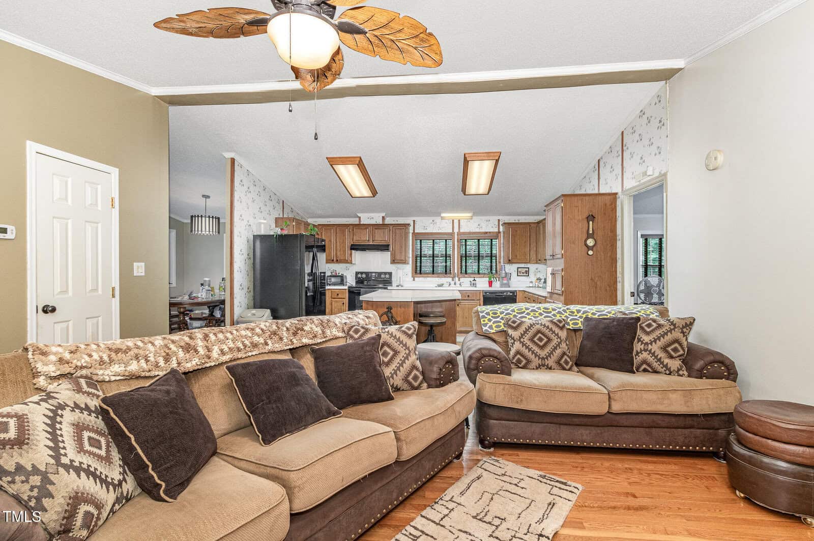 Living room with two brown sofas, patterned cushions, and a ceiling fan with leaf-shaped blades. An open view to the kitchen shows a black fridge, wooden cabinets, and a breakfast bar with stools. Walls have floral wallpaper. Wood flooring is visible.
