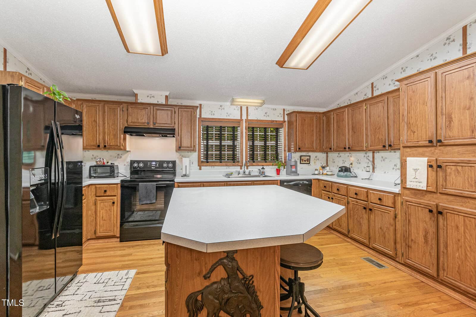 A kitchen with wooden cabinets and a central island with a decorative horse carving. Black appliances are present, including a refrigerator and stove. The walls have a floral wallpaper, and the ceiling has fluorescent lighting.