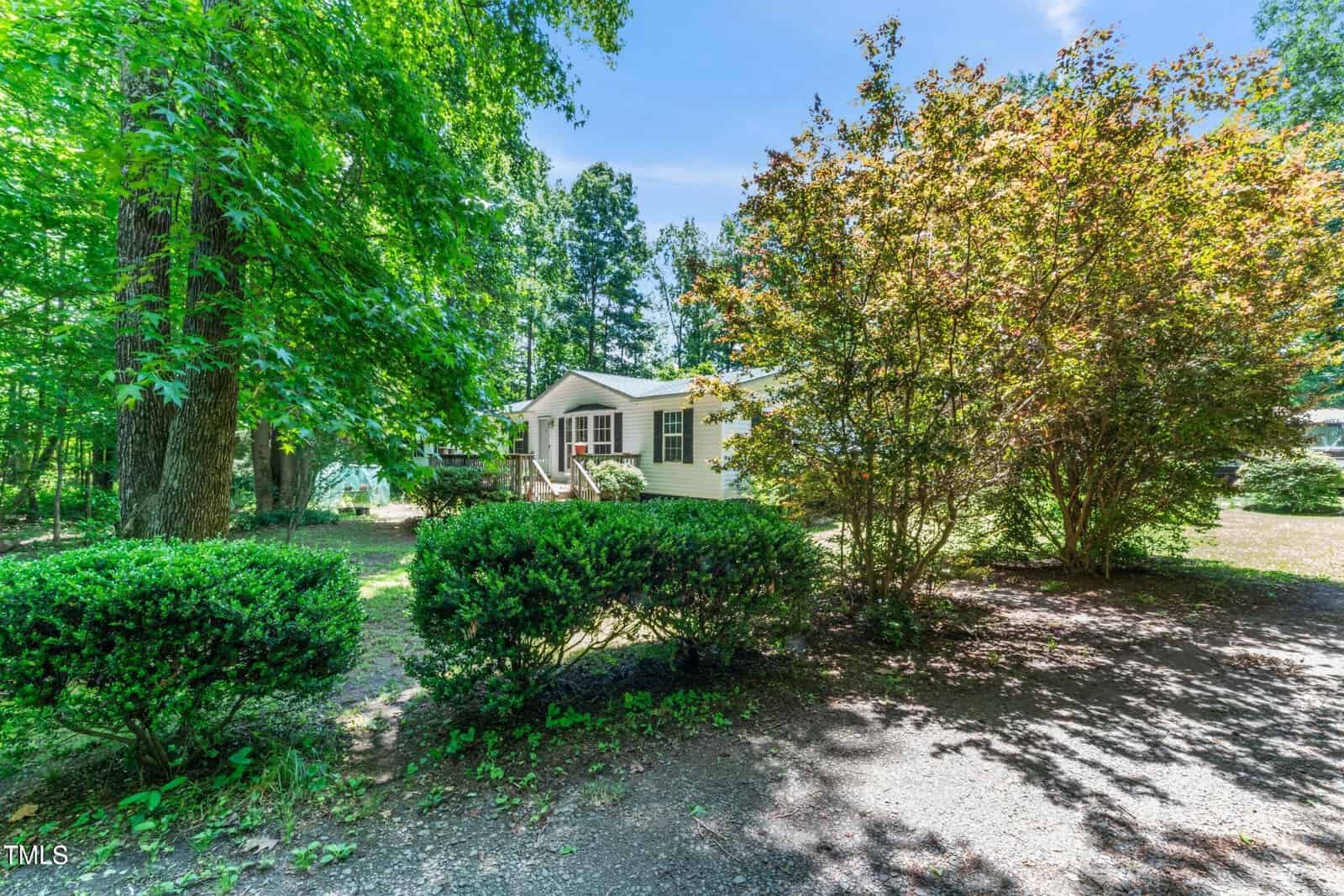 Nestled in a wooded setting at 6592 Pebble Lane, Oxford, a white house with green shutters peeks through the trees and bushes. A gravel path meanders to the entrance under a clear blue sky.