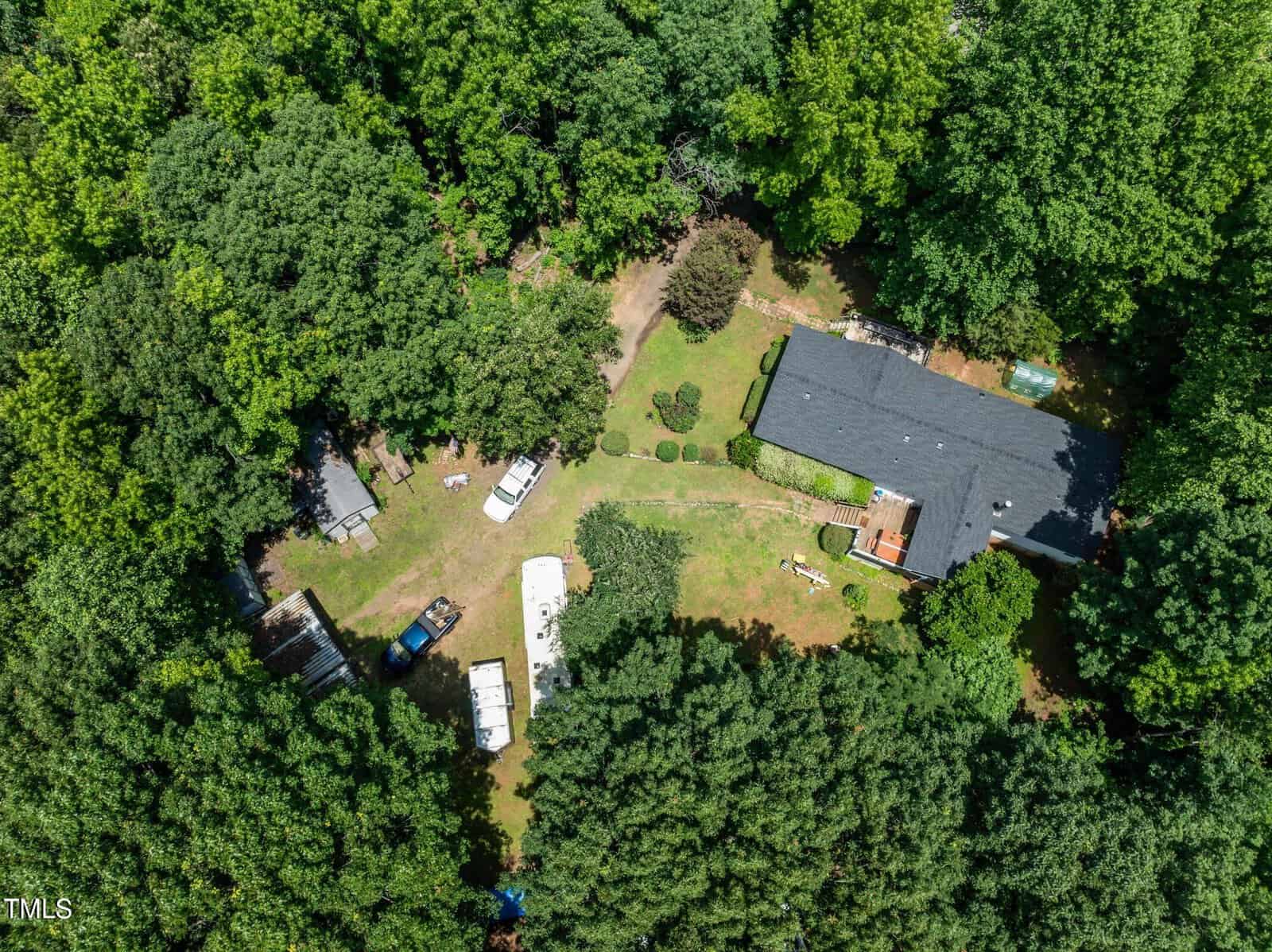 Aerial view of a house surrounded by dense green trees. The property includes a black-roofed building, vehicles, and assorted outdoor items. A grass-covered yard is visible, along with some shrubs and pathways.