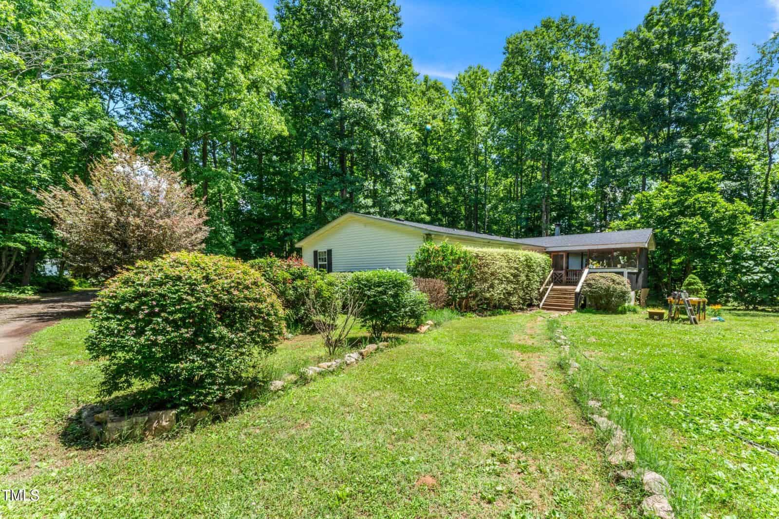 A white, single-story house is set among lush greenery, surrounded by trees. A small porch with steps leads to the entry. The lawn is well-maintained with manicured bushes and a stone path. The sky is clear and blue.