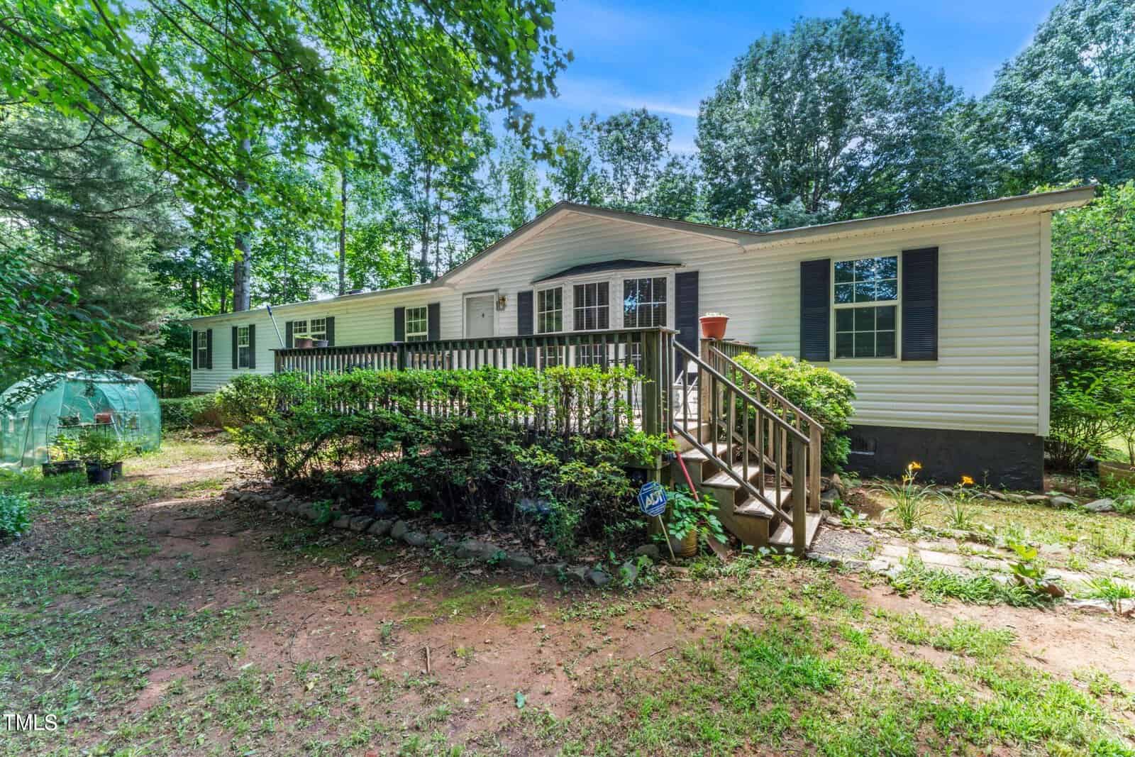 A white single-story mobile home with a wooden front porch, surrounded by trees and greenery. A small greenhouse is visible on the left side. The yard is grassy with some landscaping, and the sky is clear and blue.