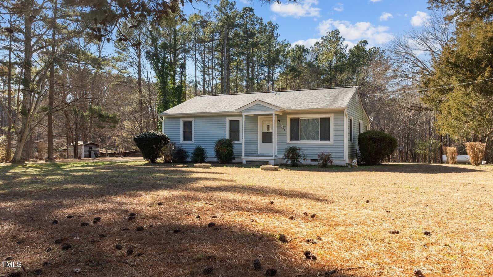A small, single-story house with light blue siding and a gray roof is situated on a plot of land with dried grass. The yard is surrounded by tall trees, and a few bushes are near the house. The sky is clear with scattered clouds.