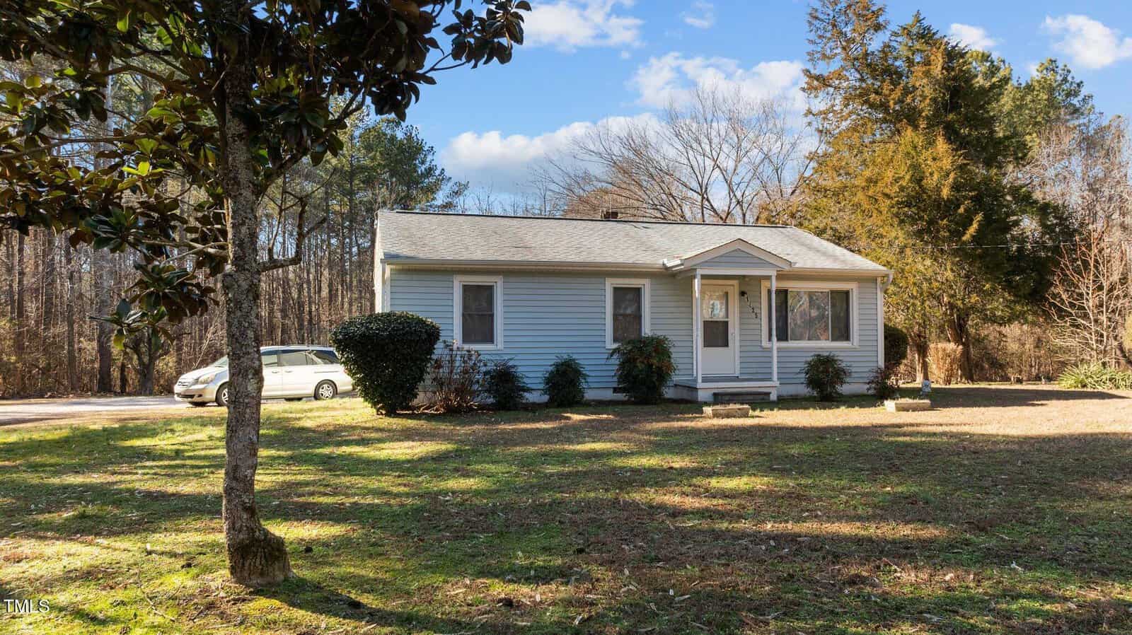 Small single-story house with light blue siding and a gray roof, set on a grassy lawn with trees and shrubs surrounding it. A white car is parked on a paved driveway to the left. The background features tall, leafless trees under a partly cloudy sky.