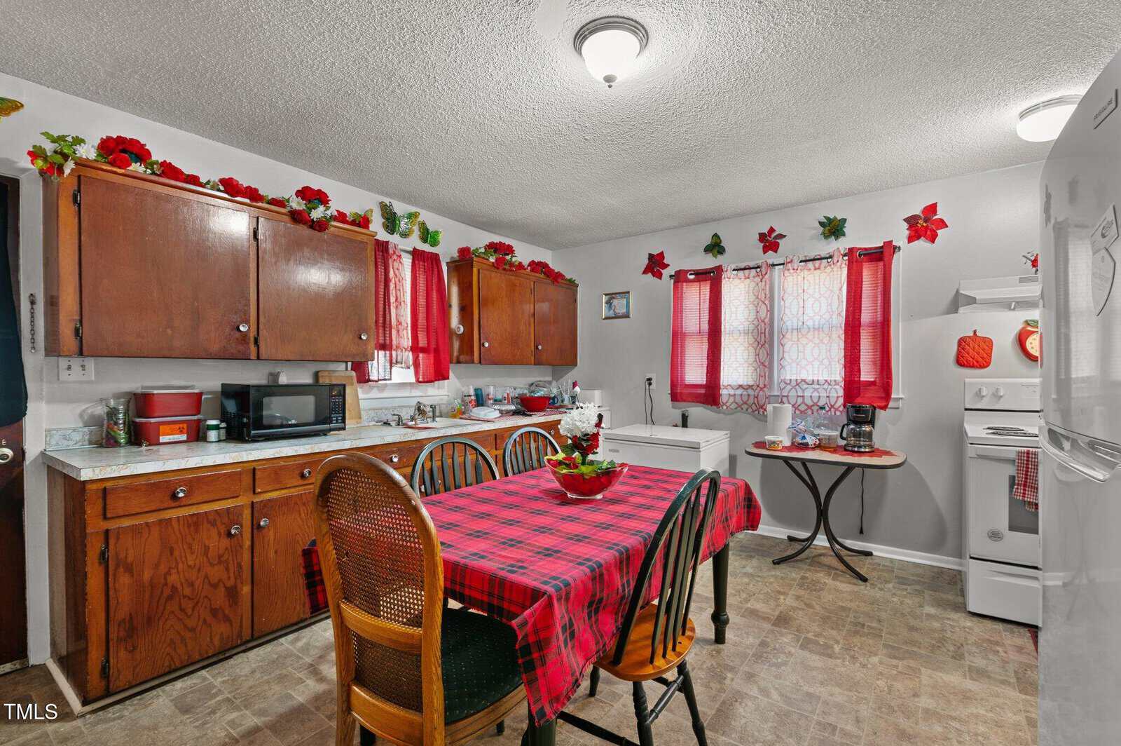 A kitchen with wooden cabinets, white appliances, and a table with a red and black checkered tablecloth. Windows have red curtains, and festive decorations are displayed above cabinets. A small table holds a coffee maker near the stove.