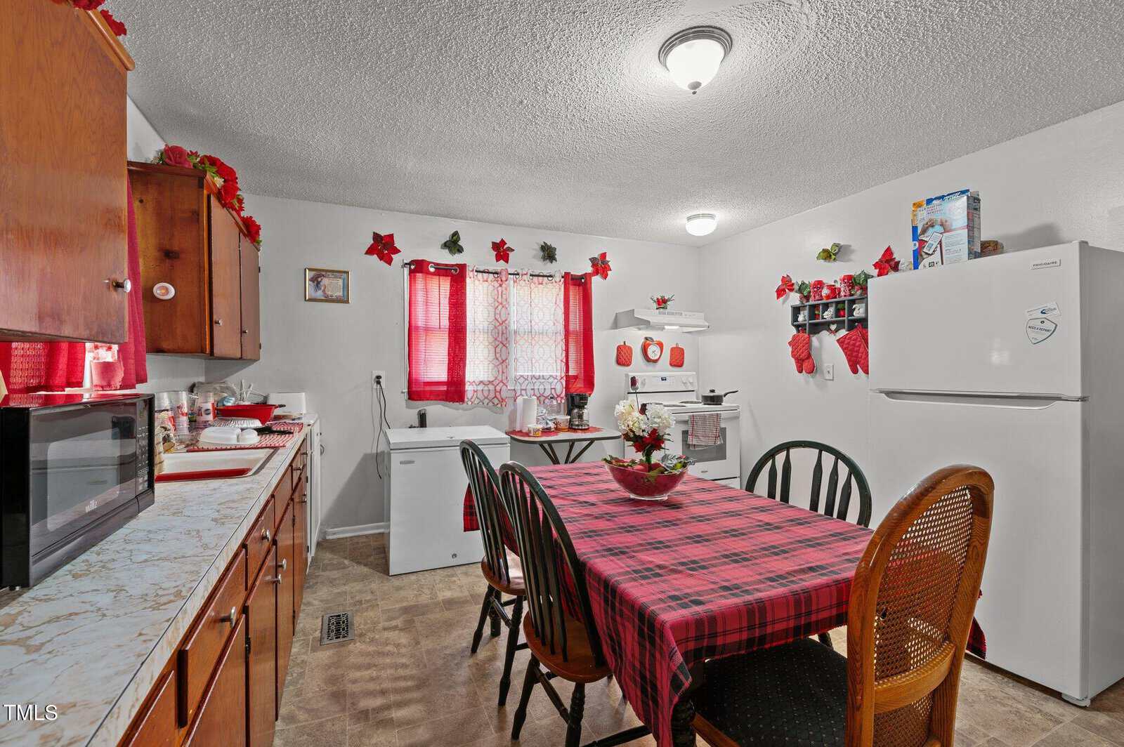 Kitchen with a plaid tablecloth on a rectangular table surrounded by chairs. Wooden cabinets line the wall to the left, a refrigerator to the right. A window with red curtains, stove, and small decor items are visible. Ceiling light illuminates the room.