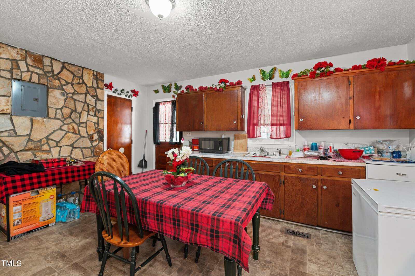 A kitchen with wooden cabinets, a stone accent wall, and red plaid tablecloth on a dining table with four chairs. Red decorations and butterfly accents line the cabinets. A microwave, dishwasher, and cleaning supplies are visible.