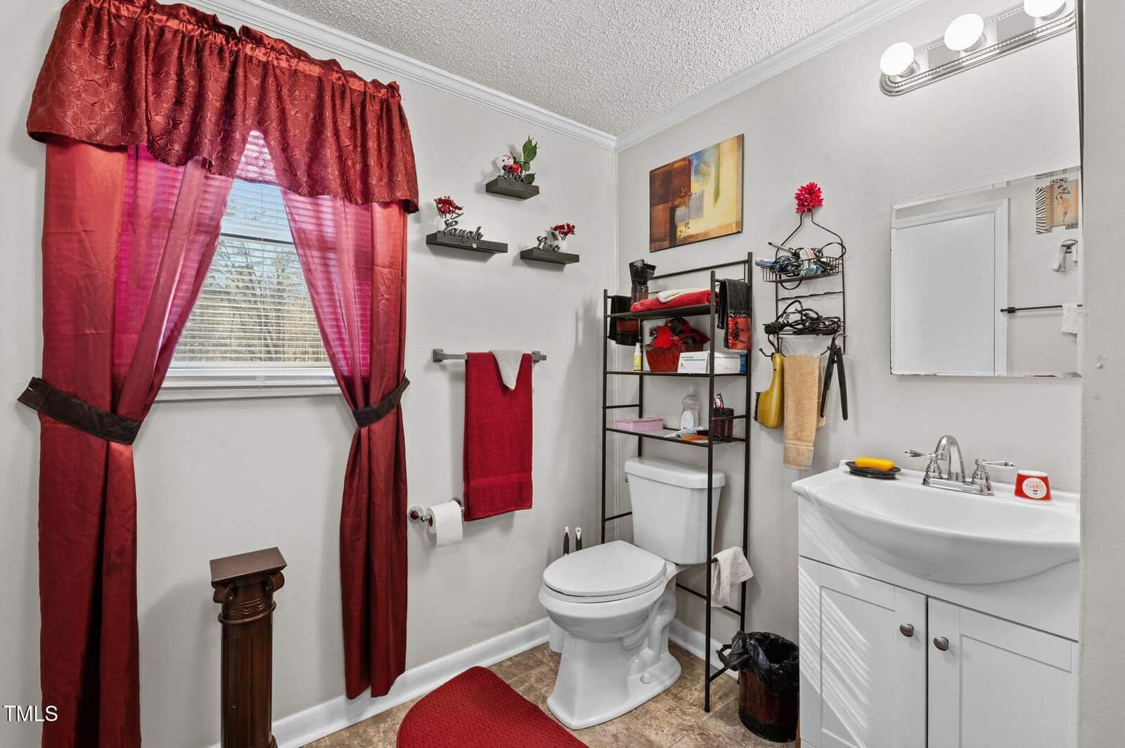 A small bathroom with a white sink and toilet. Red towels, curtain, and rug provide accents. Shelves hold decor, including red flowers and candles. A window with a red curtain is on the left. Walls are painted light gray, and a small artwork is hung.