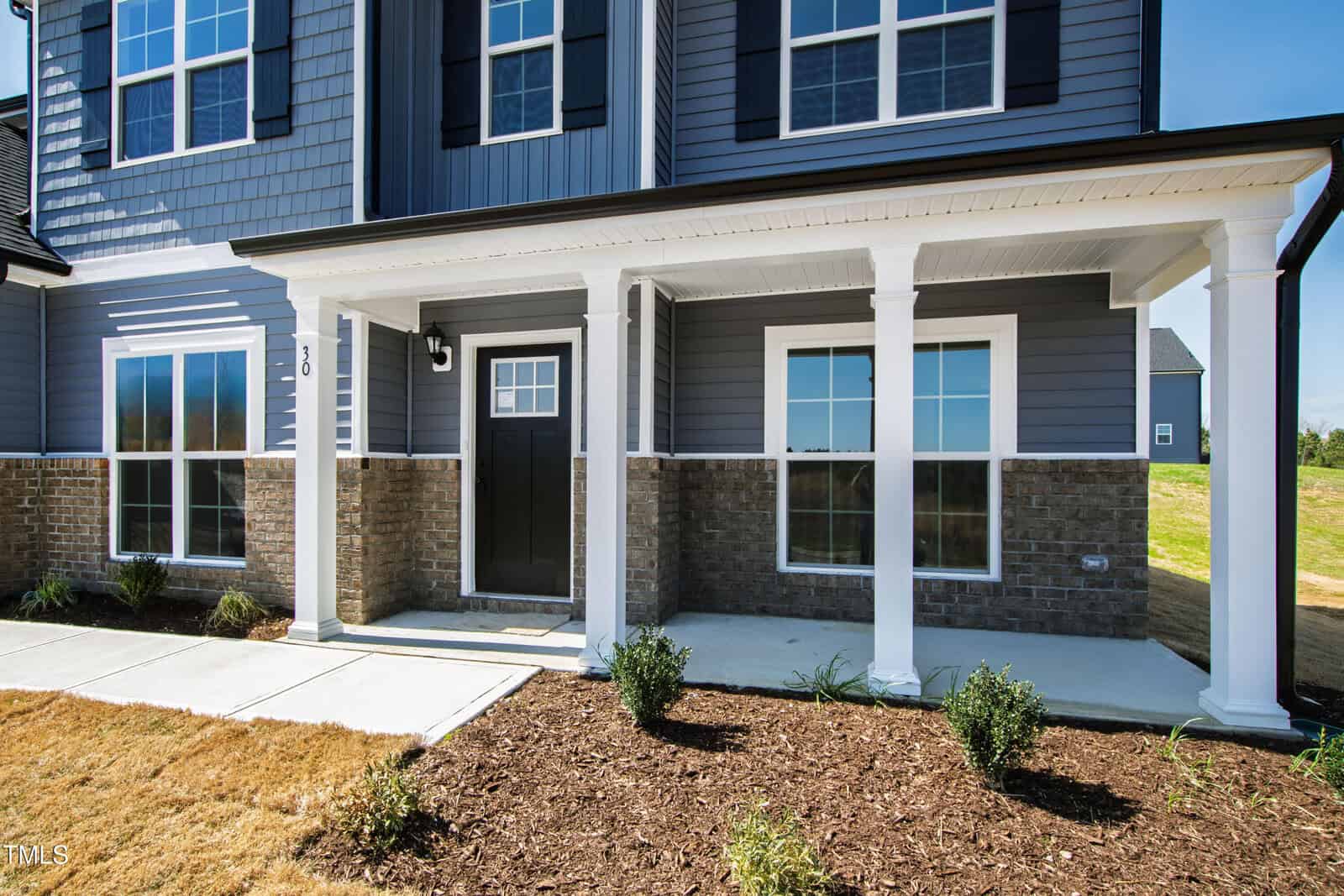 A modern two-story house with gray siding, white trim, brick lower facade, a covered front porch with white columns, black front door, and landscaped mulch bed with small plants.
