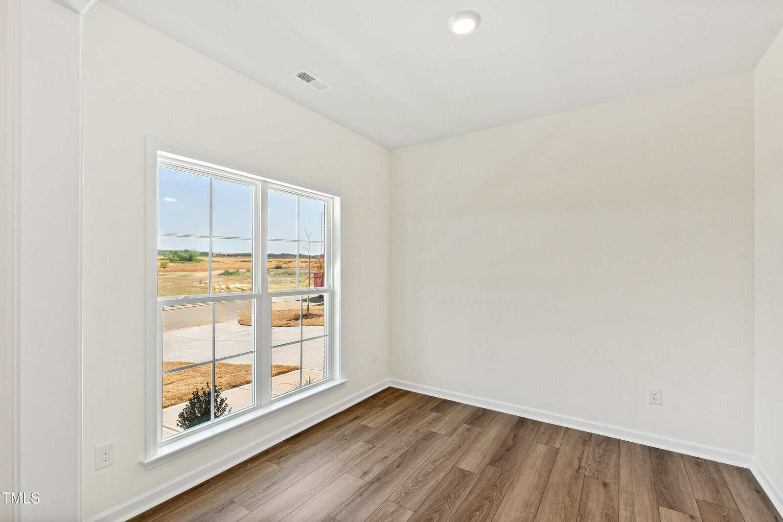 A small, empty room with white walls, a wood floor, and a large window showing a view of a street and an open landscape outside. Ceiling light is turned on.