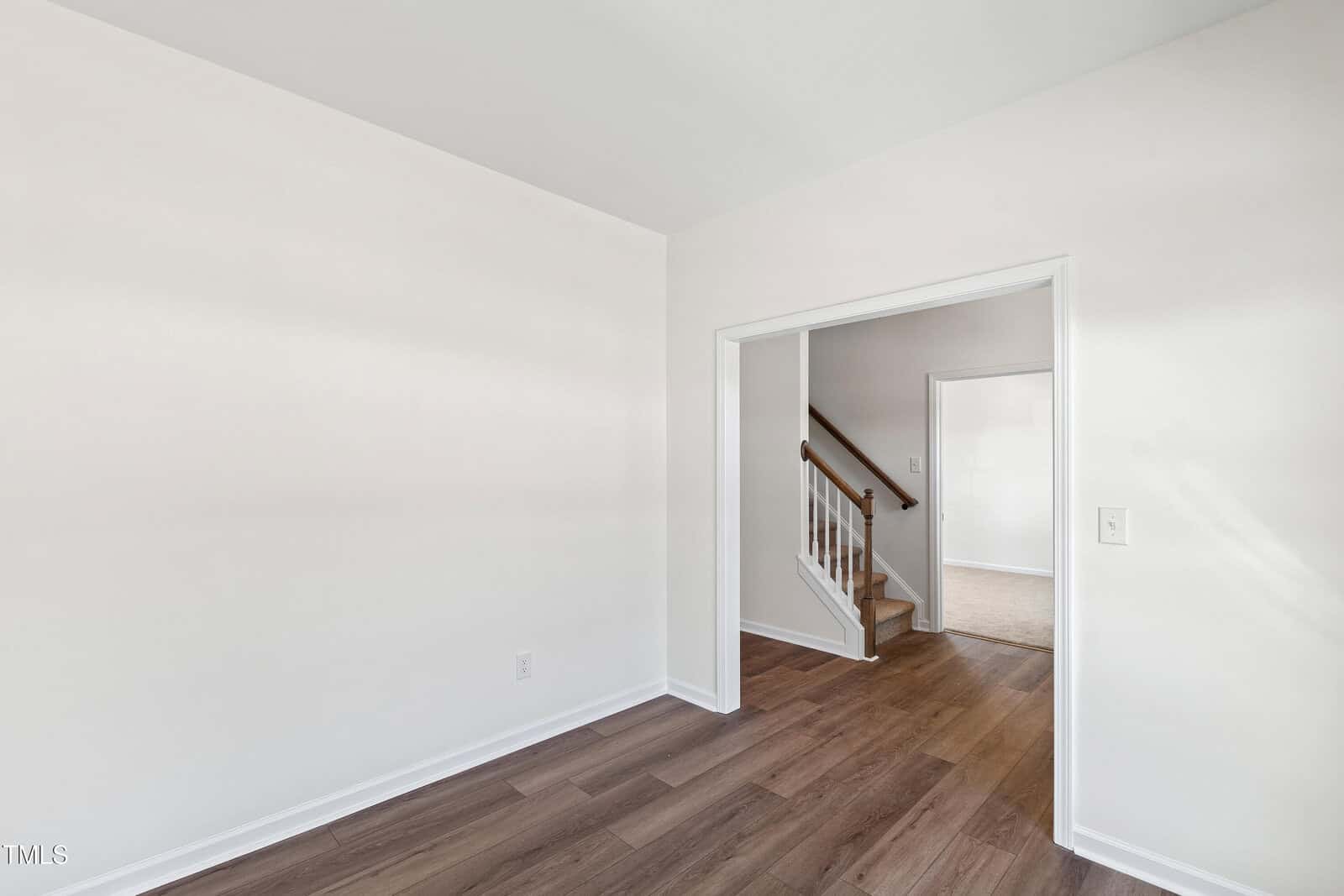 Room with white walls, wood-look flooring, and a doorway leading to a stairway with a white railing and carpeted steps. Natural light brightens the space. No furniture is present.