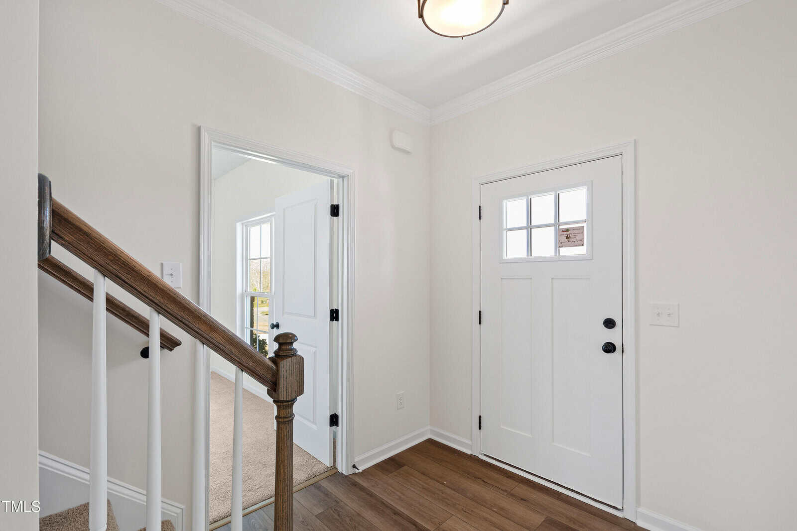 A small entryway with a white front door featuring a window, a staircase with wood steps and white railing, and an open doorway leading to a carpeted room with a window. Walls are painted light beige.