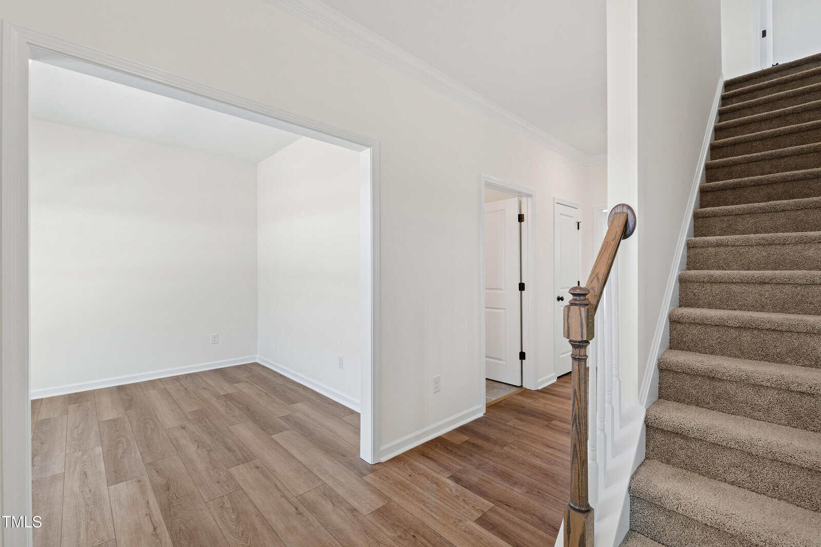 A hallway with wood flooring leads to a carpeted staircase on the right. There is an open doorway to a small room on the left and two closed white doors further down the hall. Walls are painted white.