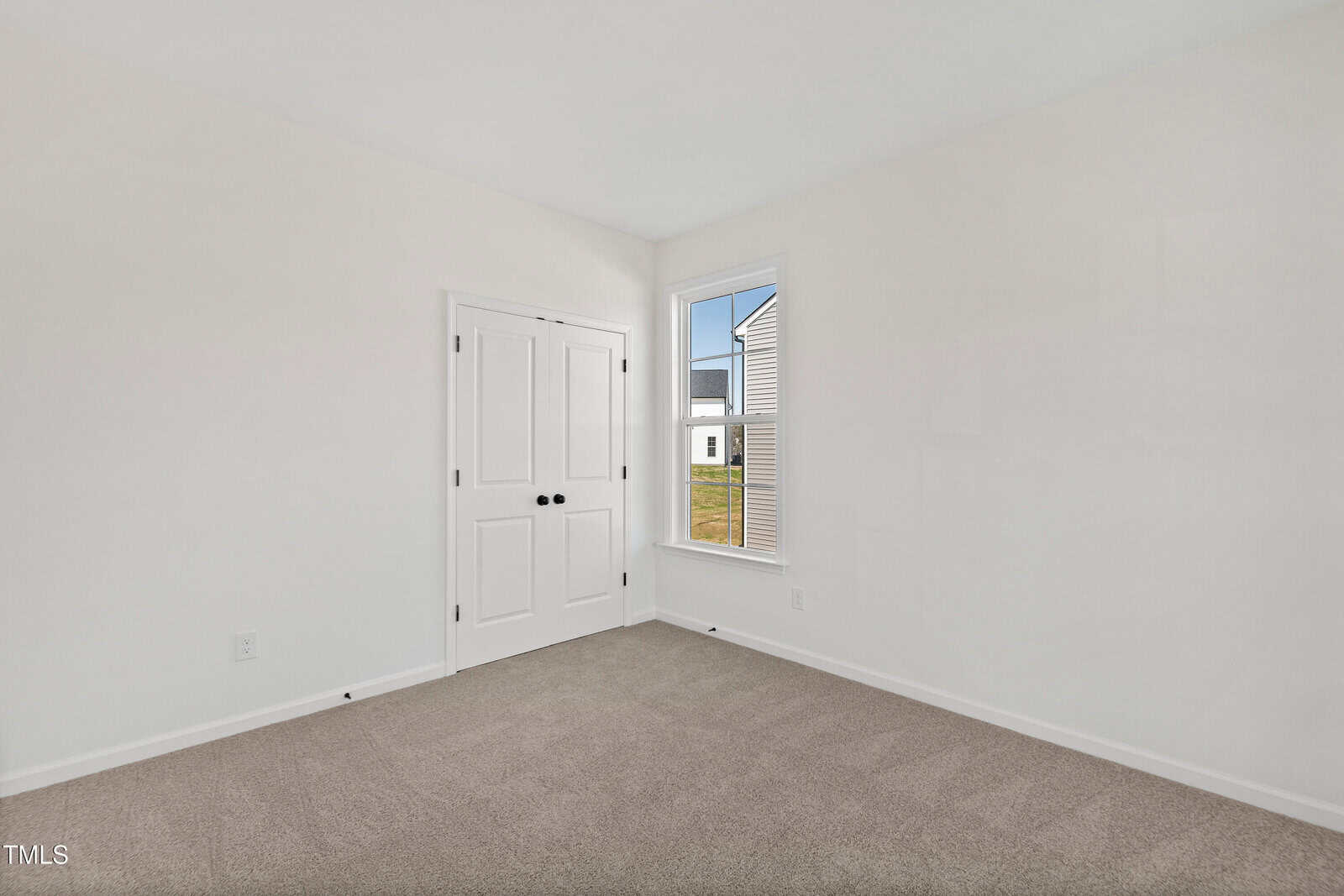 Empty room with beige carpet, white walls, a double-door closet with black handles, and a single window showing an exterior view of a house and grass. The room is well-lit with natural light.