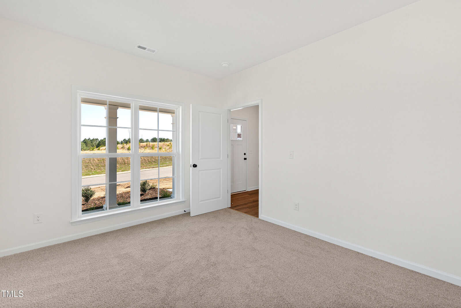 Empty room with beige carpet, white walls, a large window showing a grassy yard, and an open white door leading to a hallway with hardwood flooring. Natural light fills the space.