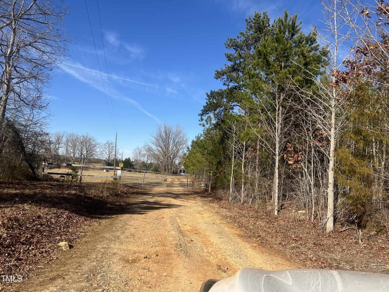 A dirt road leads to a gate in a rural area. There are bare trees on the left and pine trees on the right. The sky is clear and blue. A portion of a vehicle is visible in the foreground.