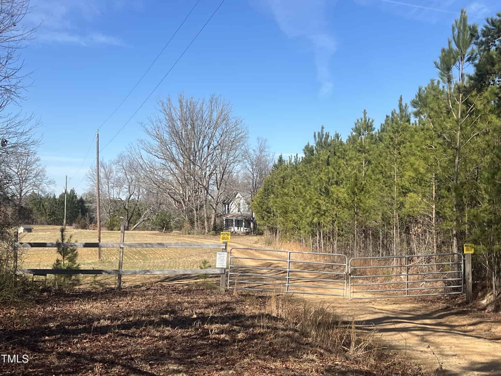 A rural scene shows a dirt driveway leading through a metal gate towards a house partially obscured by trees. The surrounding area includes leafless trees and pines. The sky is clear and blue. Signs are posted on the gate.