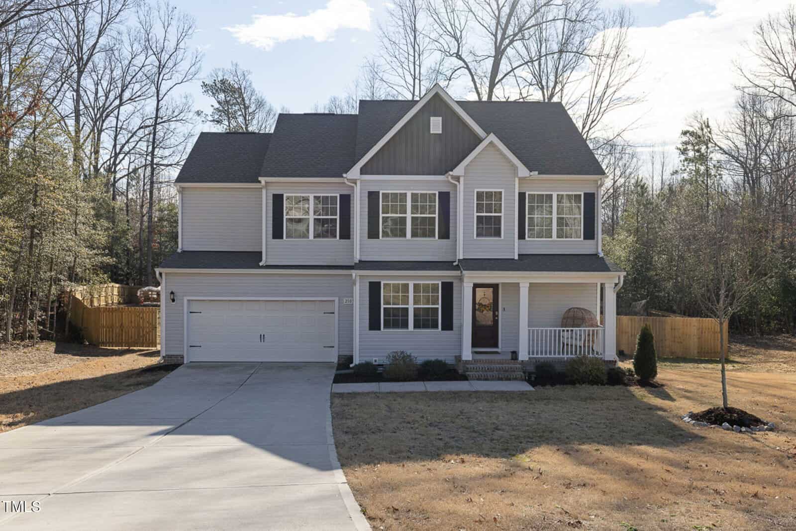 A two-story suburban house with gray siding, white trim, and a covered front porch. The home has a double garage, a large driveway, and a fenced backyard with trees and sparse grass.
