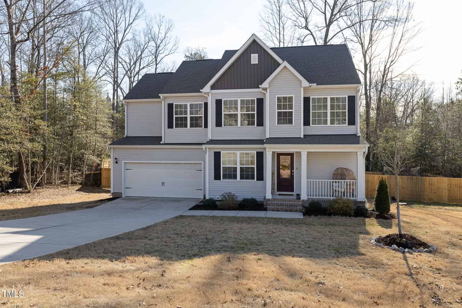 A two-story suburban house with gray siding, white trim, and a dark shingled roof. It has a double garage, front porch, and a large lawn bordered by trees and wooden fencing.