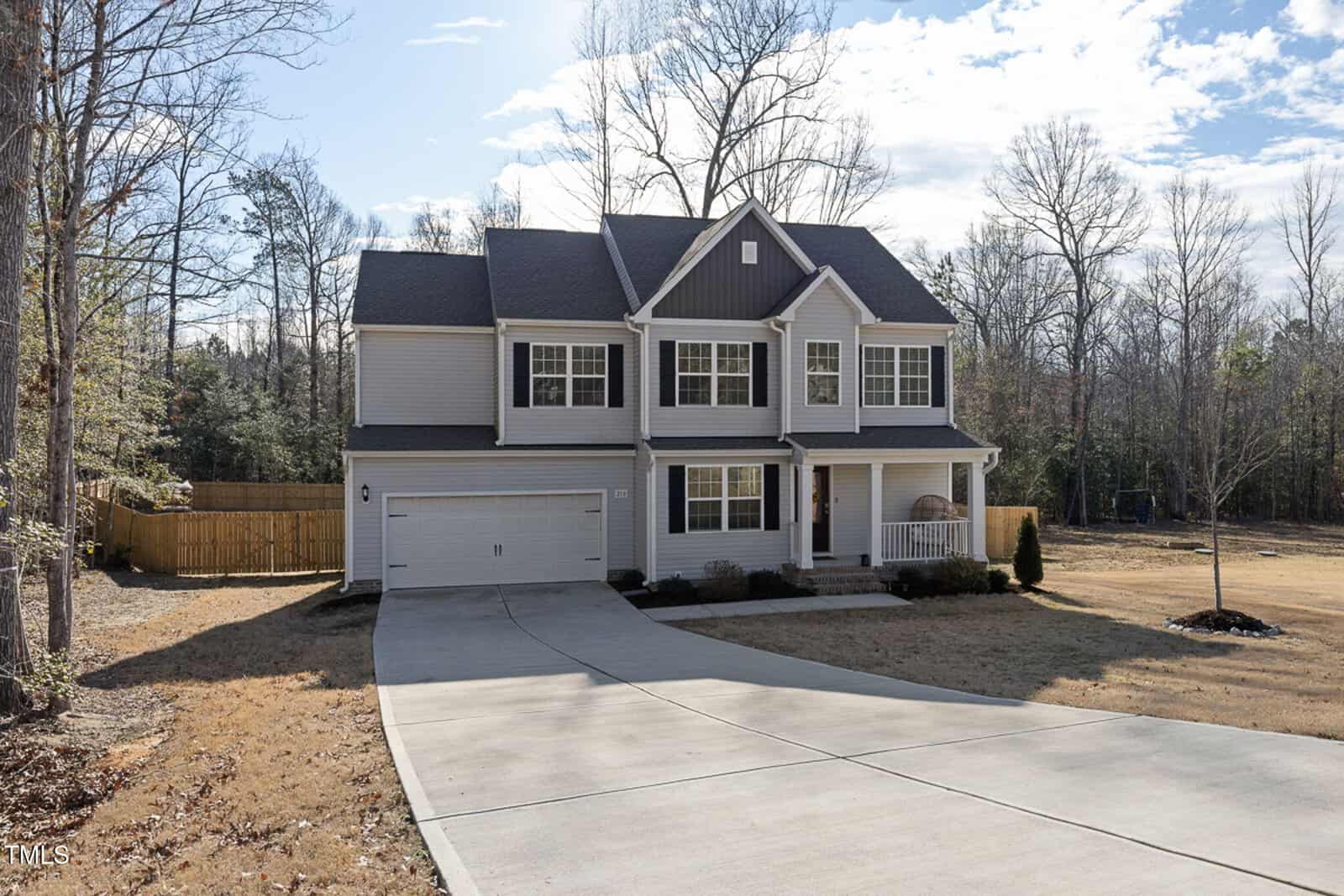A two-story suburban house with gray siding and white trim, attached two-car garage, covered front porch, and a curved concrete driveway. The yard is mostly grass with some trees and a wooden fence on the left.