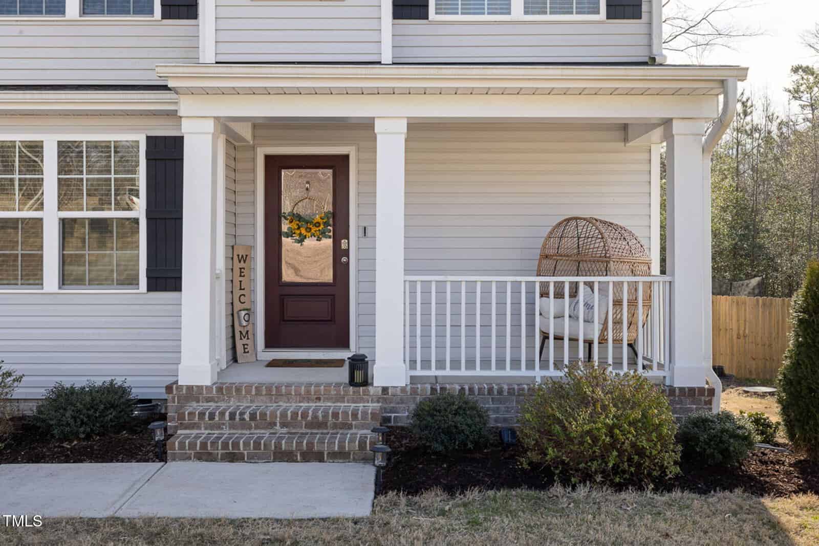 The image shows a front porch with a brick step, a brown door decorated with a wreath, a "Welcome" sign, a white chair with cushions inside a wicker egg-shaped frame, and shrubs bordering the house.