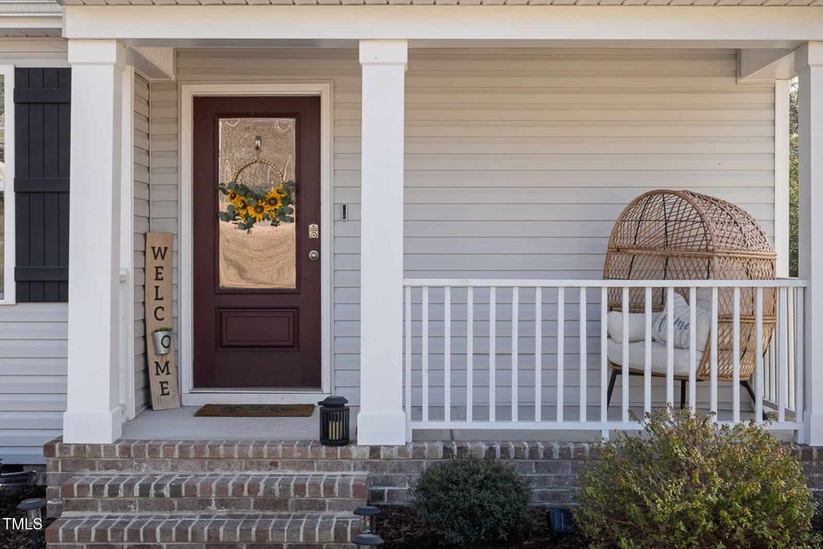 Front porch of a house with a brown door displaying a floral wreath, a vertical wooden "Welcome" sign, a wicker egg chair with cushions, brick steps, white railings, and some small shrubs in front.