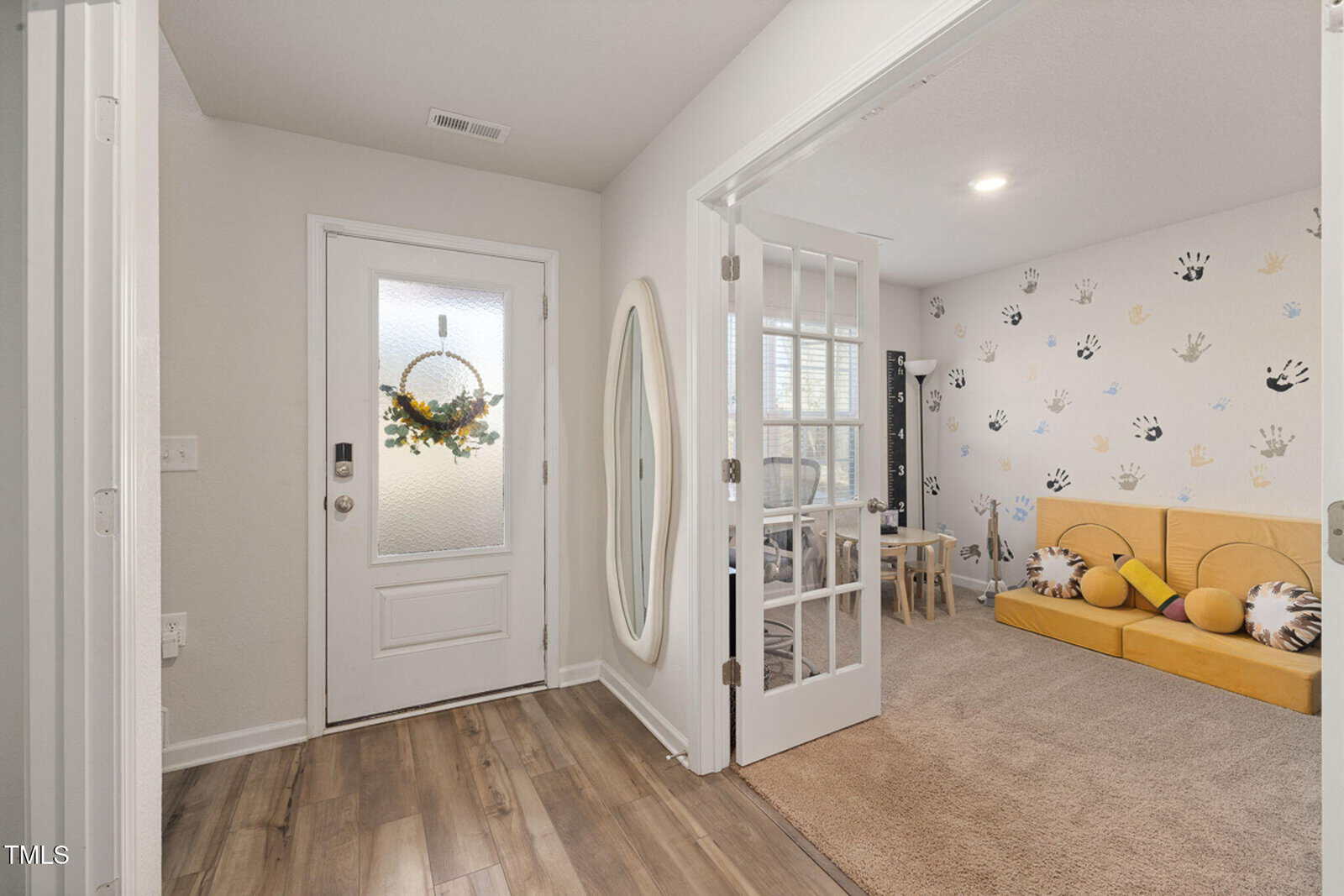 A hallway with a white front door, oval mirror, and wood floors. To the right, a room is visible through glass french doors, featuring a yellow sofa, a small table, and a wall decorated with colorful handprints.