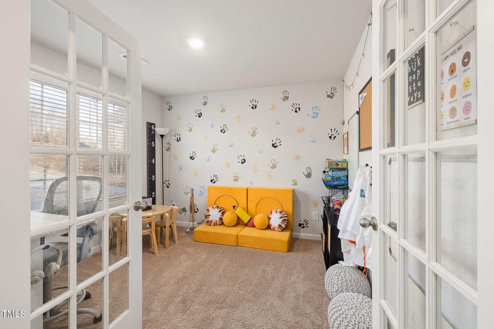 A children’s playroom with a yellow sofa, small table and chairs, wall decorated with colorful handprints, shelves with toys, and a coat rack. The room has carpeted flooring and French doors with glass panels.