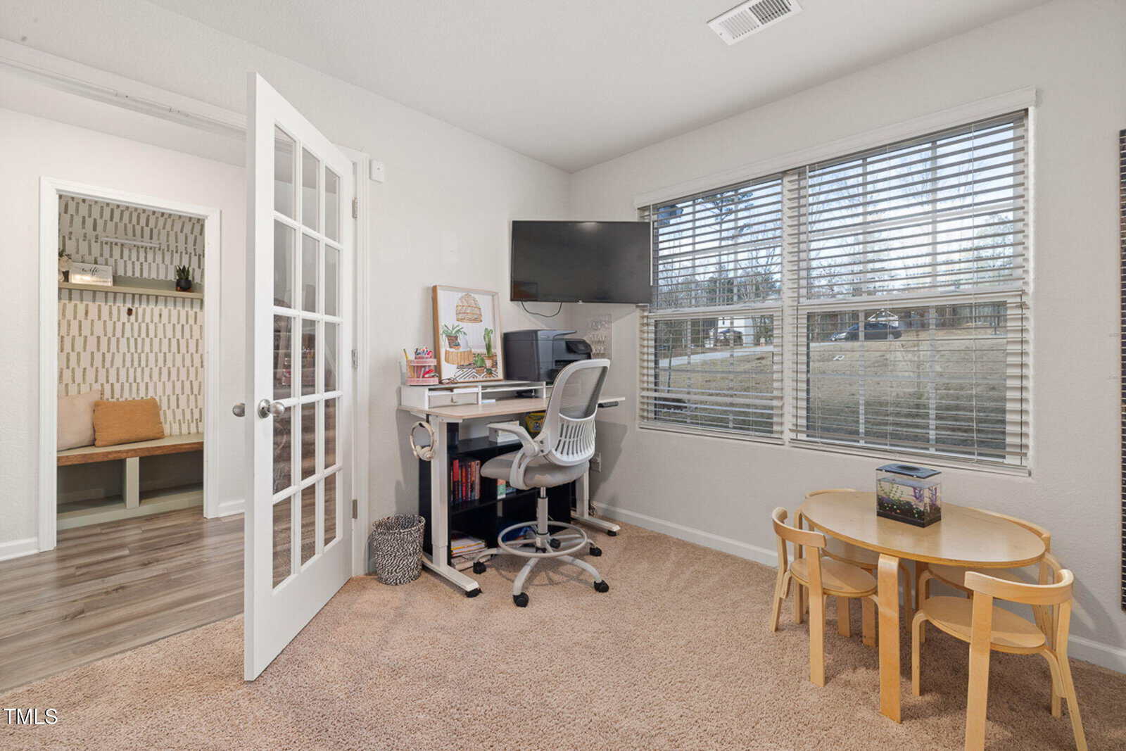 A home office with a desk, chair, computer, and monitor near a window. A small kids’ table with chairs and a fish tank is in the corner. An open door leads to a hallway with shelves and a bench.
