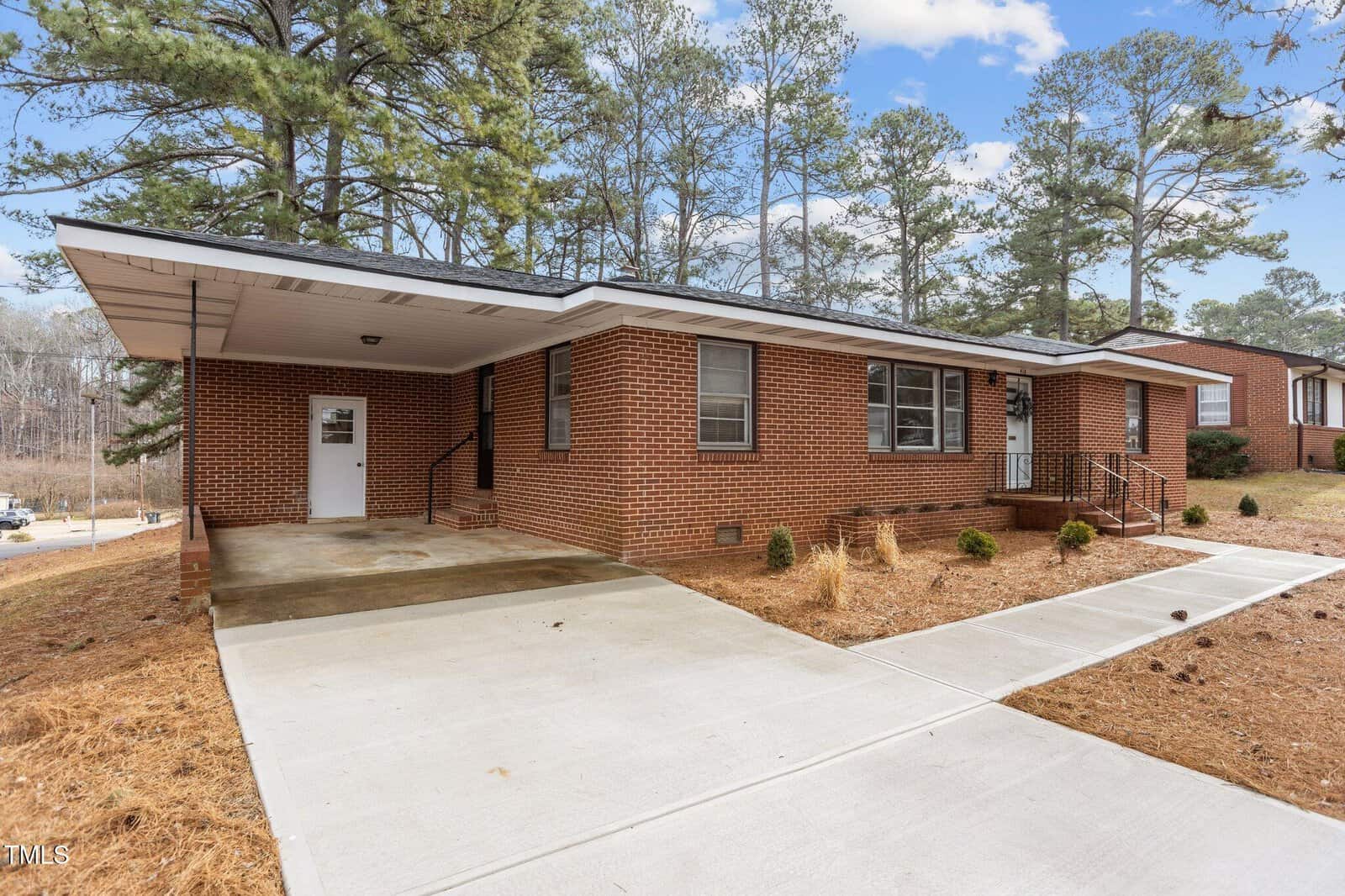 A single-story red brick house with a covered carport, concrete driveway, and walkway, surrounded by pine trees and minimal landscaping. The house has several windows and white trim.