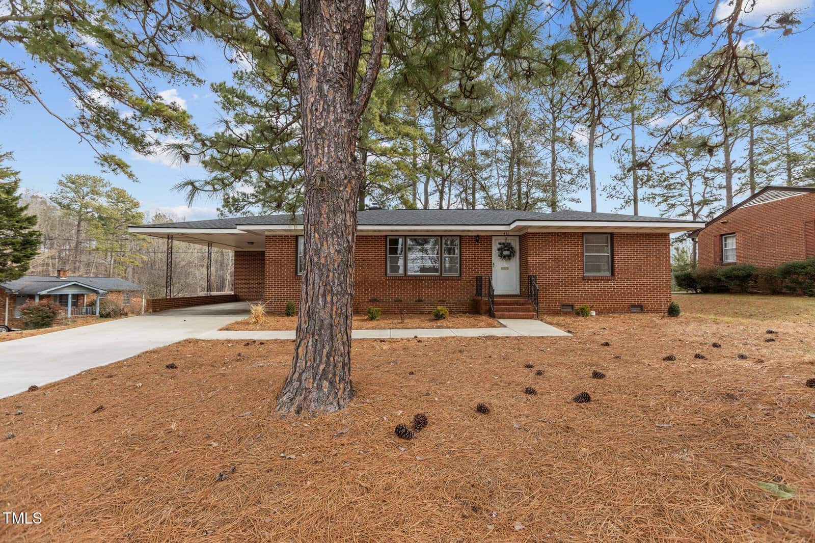 A single-story brick house with a covered carport on the left side, a paved driveway, and a large pine tree in the front yard. The ground is covered with pine needles and there are small shrubs near the house.