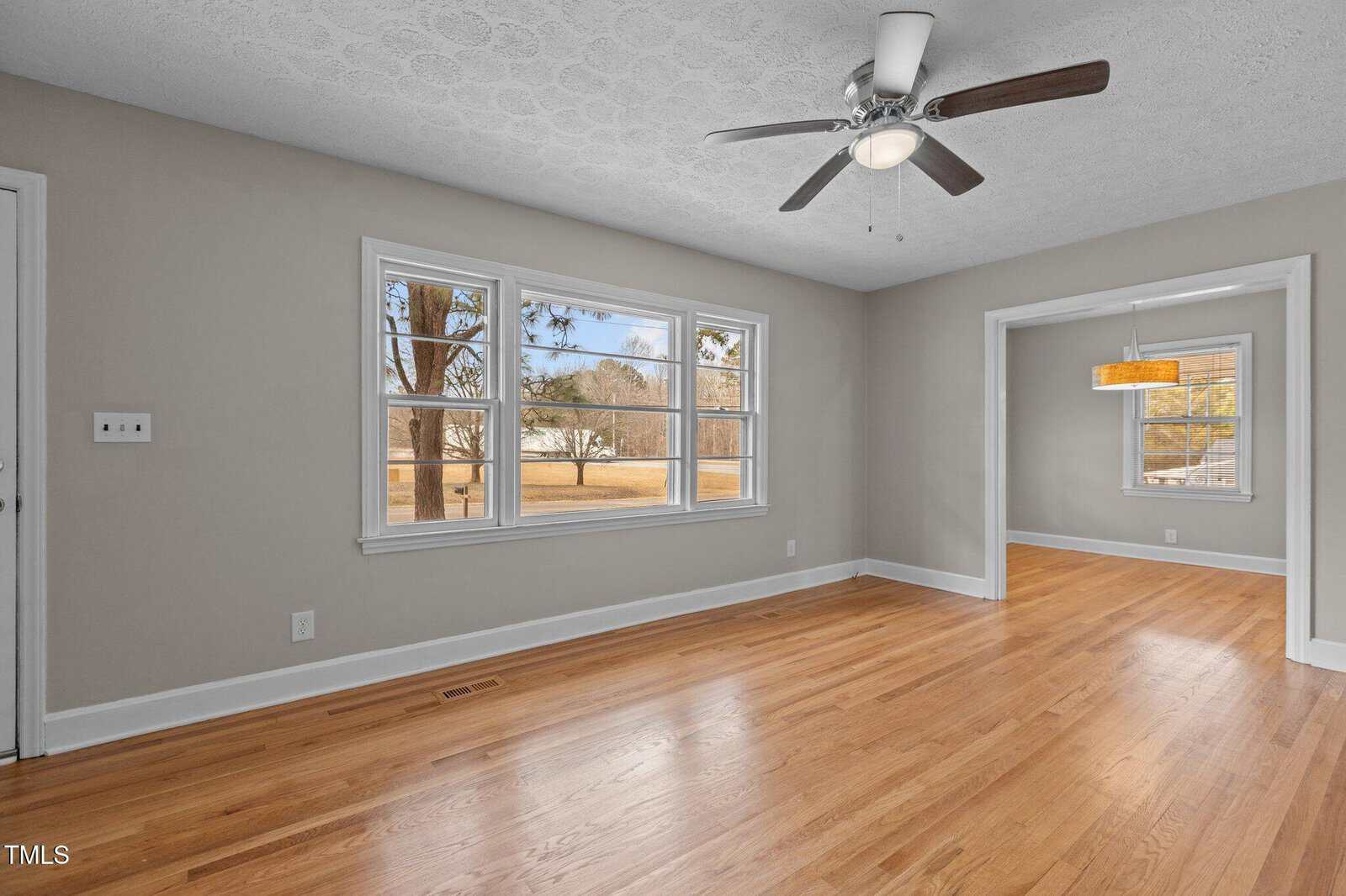 A bright, empty room with light wood floors, beige walls, a ceiling fan, three large windows, and an open doorway leading to another room with similar flooring and a hanging light fixture.