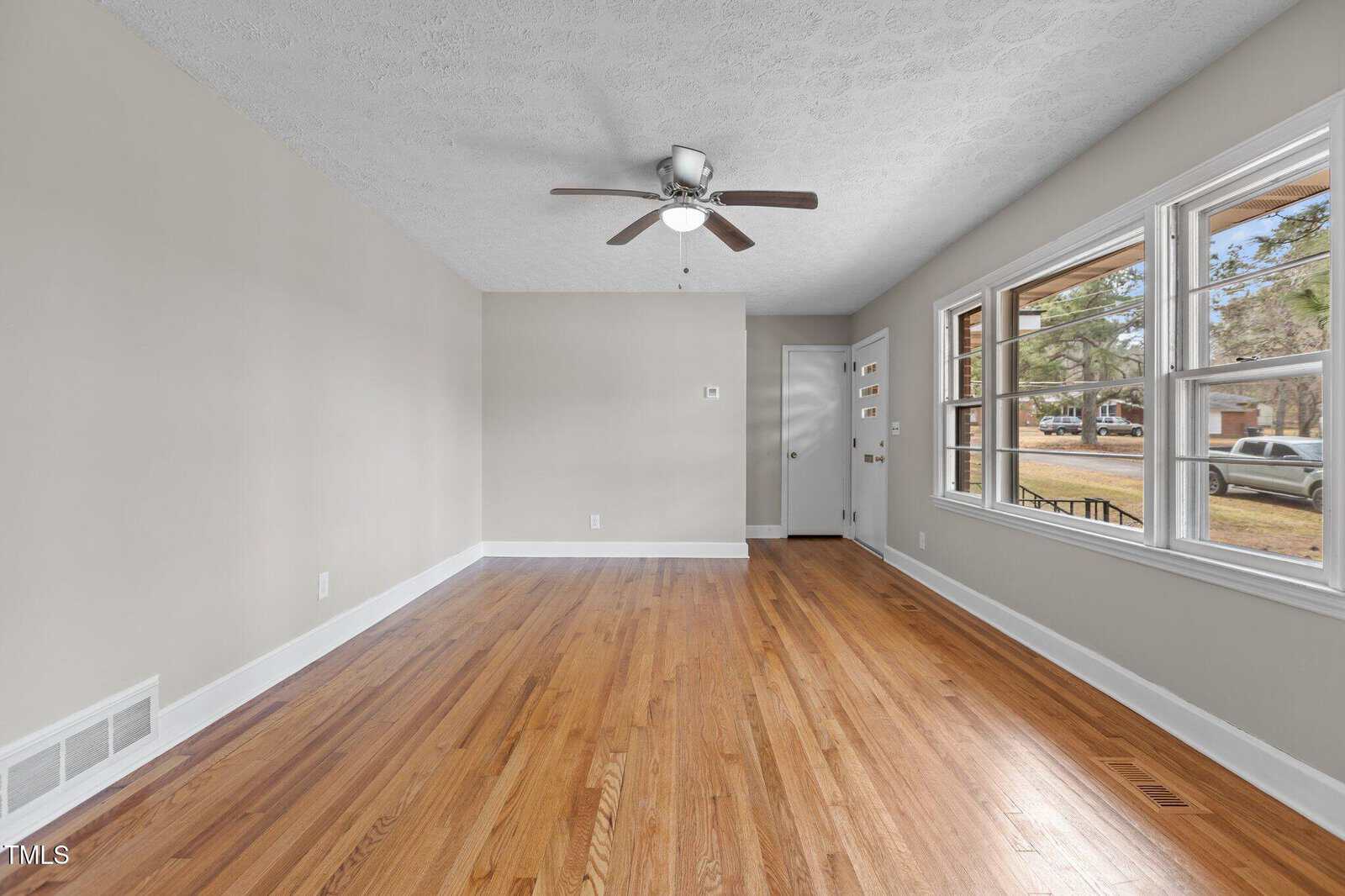 Unfurnished living room with light beige walls, hardwood floors, a ceiling fan with a light, and several windows. The entry door and a closet door are visible on the far wall. Natural light comes through the windows.