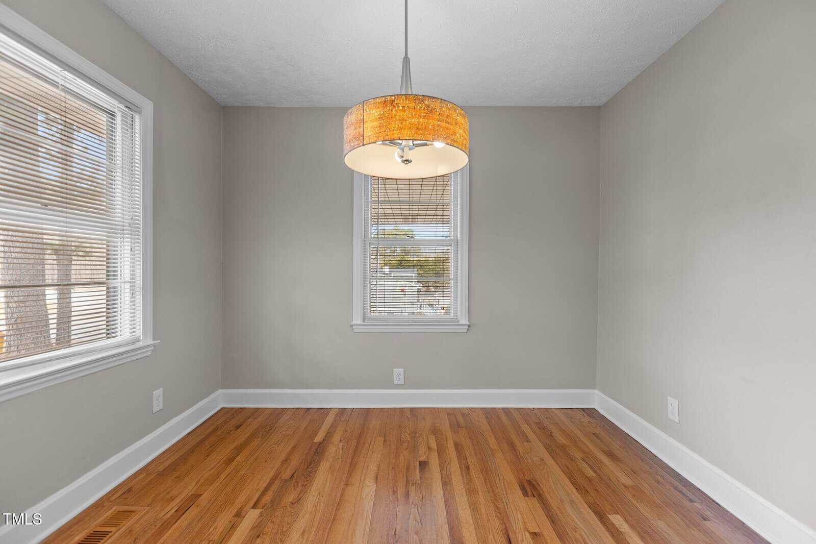 An empty room with light beige walls, a hardwood floor, two windows with blinds, white trim, and a single ceiling light fixture with a woven shade.