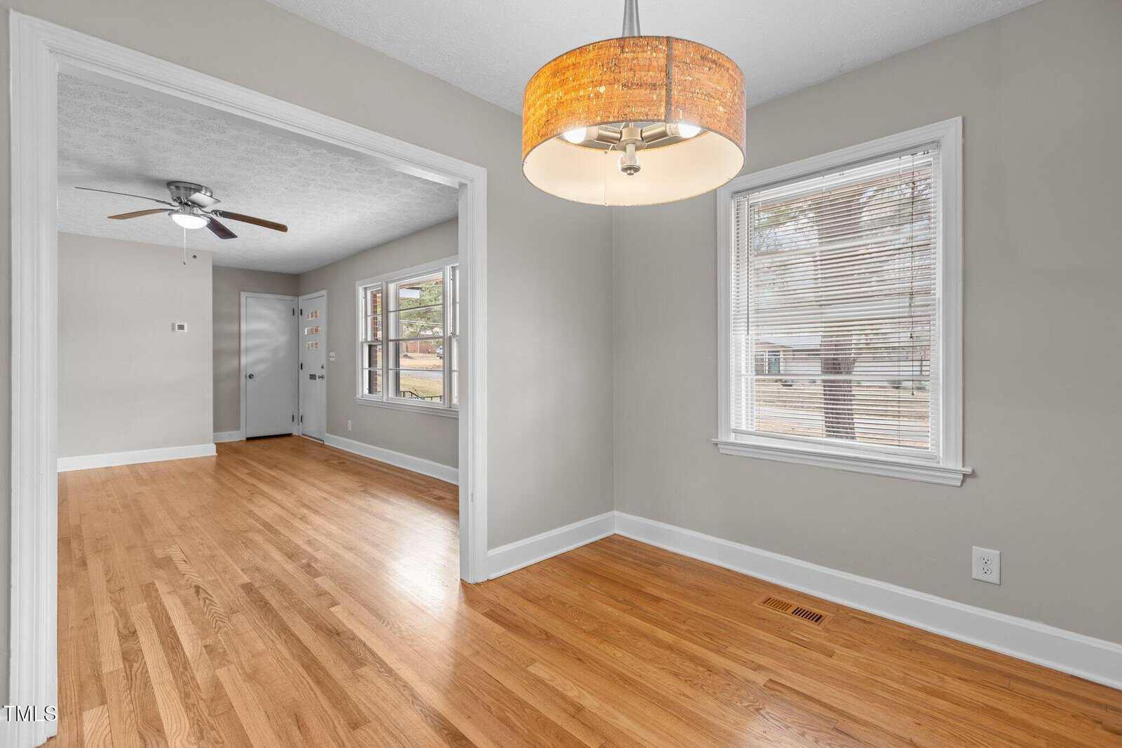A dining area with hardwood floors, a large window with blinds, and a round ceiling light fixture. An adjacent room features similar flooring, two windows, and a ceiling fan with light. Walls are painted light gray.