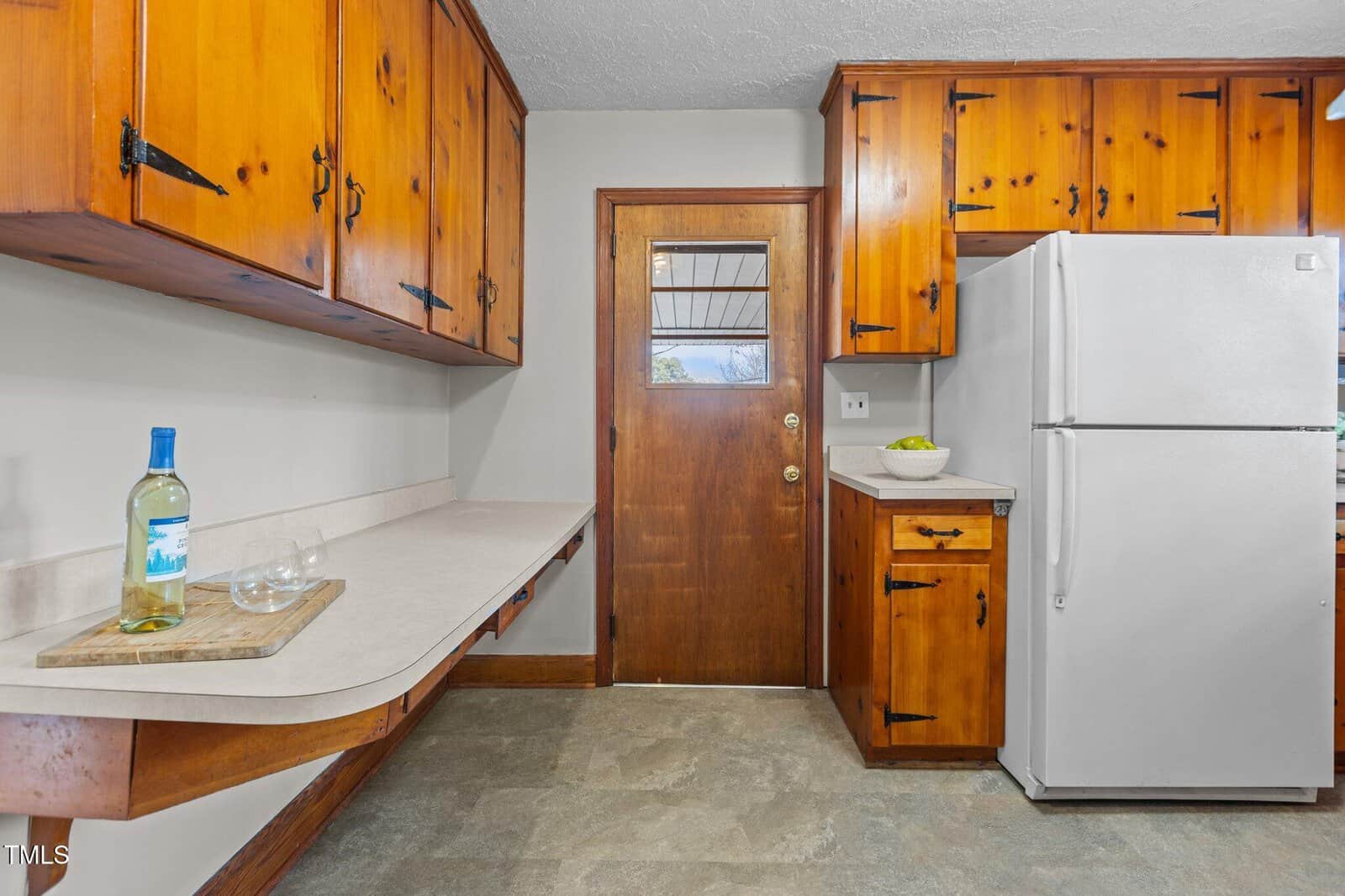 A kitchen with wooden cabinets, a white refrigerator, a wooden door with a window, and a countertop holding a wine bottle, two glasses, and a bowl of apples. The floor appears to be stone or tile.