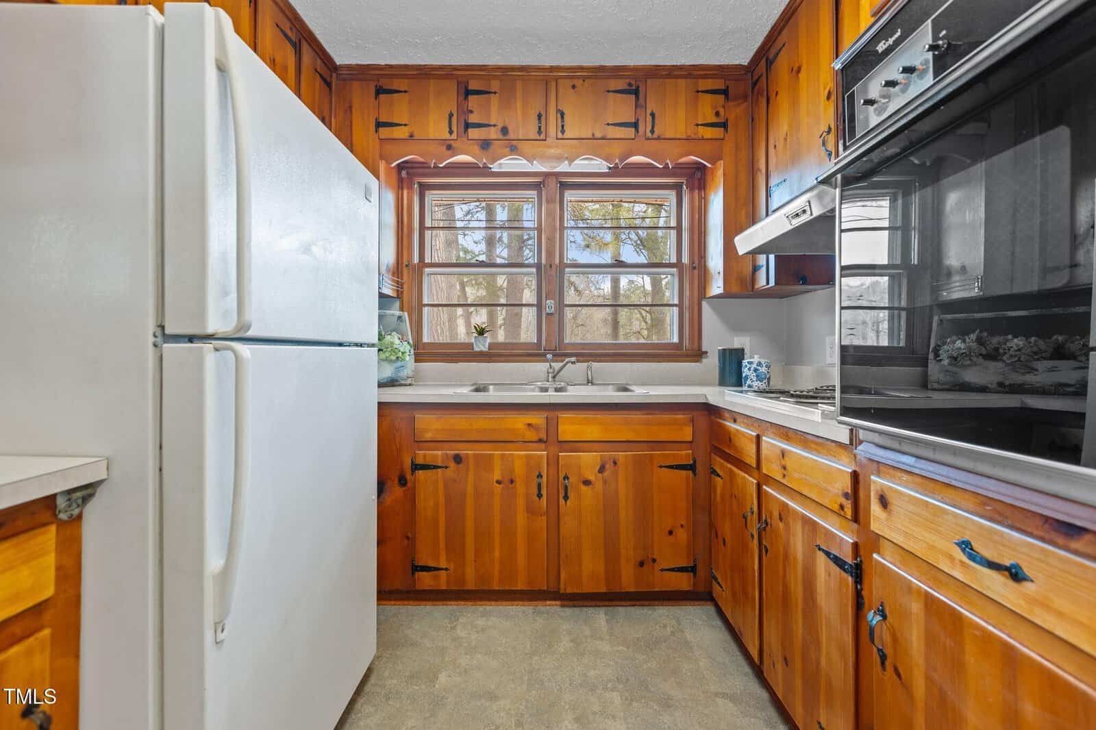 Kitchen with wood cabinets, white refrigerator, dual sink under a window, and built-in black oven. Countertops are light-colored, and natural light enters through a three-panel window above the sink.