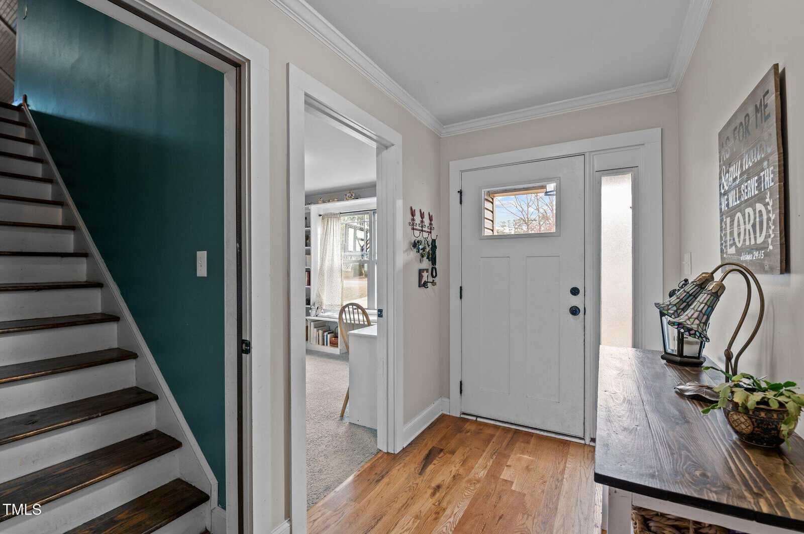 A hallway with wooden floors, white walls, and crown molding. A staircase with dark wooden steps is on the left, a table with decor is on the right, and a white front door with glass panels is straight ahead.