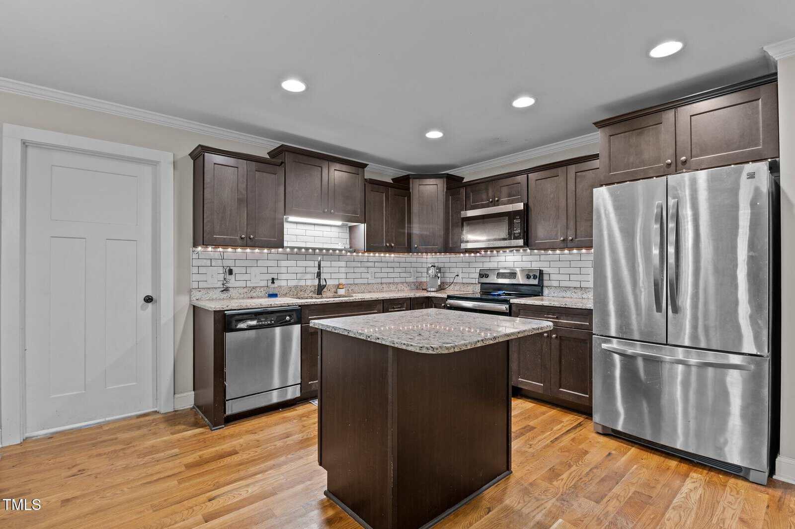 Modern kitchen with dark wood cabinets, stainless steel appliances, an island with granite countertop, subway tile backsplash, recessed lighting, and hardwood floors. A white door is on the left wall.