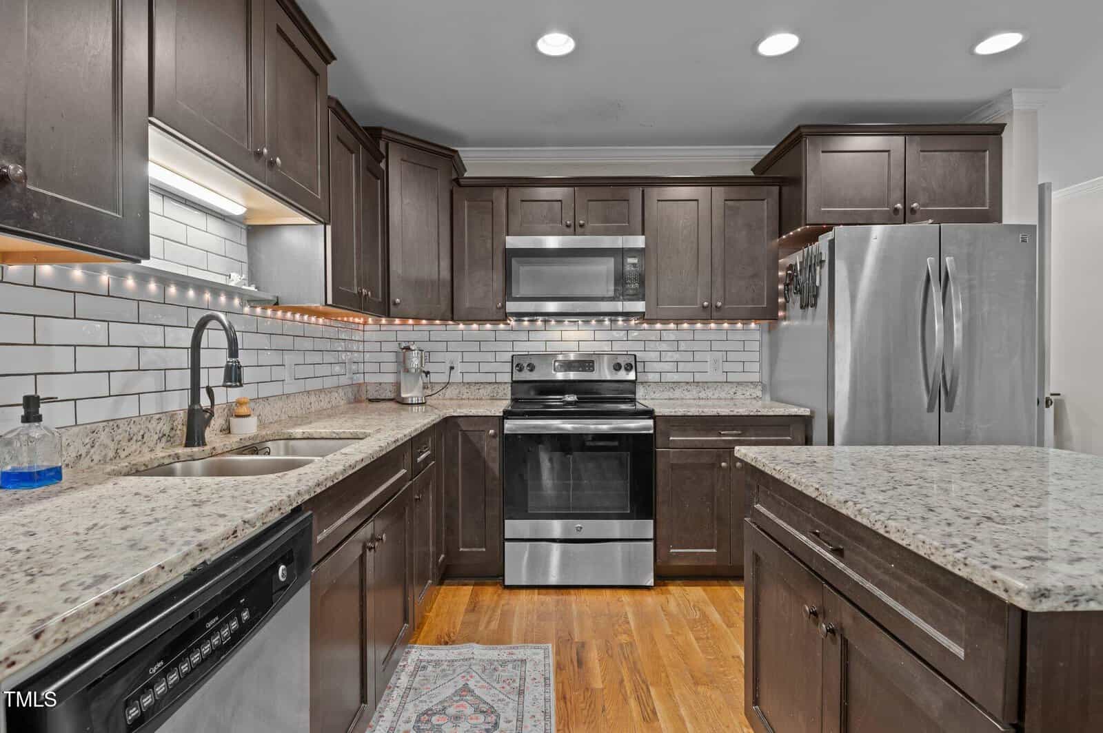 A modern kitchen with dark wood cabinets, stainless steel appliances, granite countertops, white subway tile backsplash, built-in lighting under cabinets, and a patterned rug on a wooden floor.