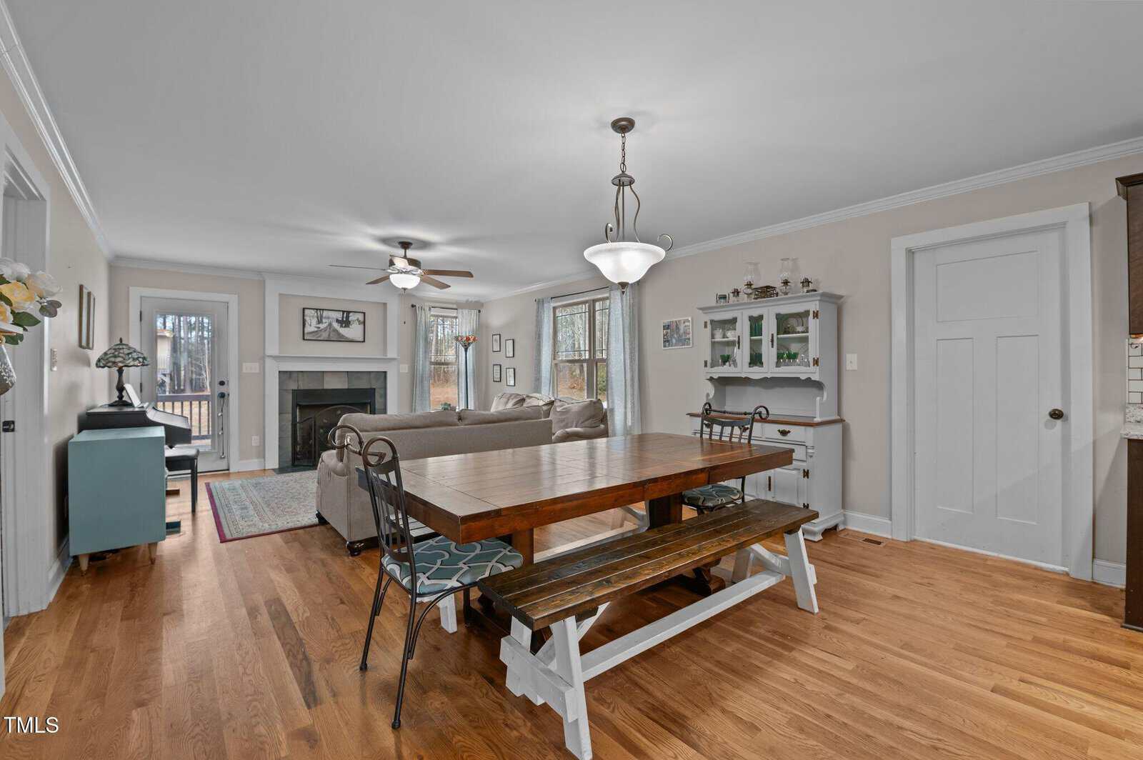 Open-plan living and dining area with wooden floors, a rectangular dining table with benches, a sofa facing a fireplace, ceiling fans, a light fixture above the table, and windows letting in natural light.